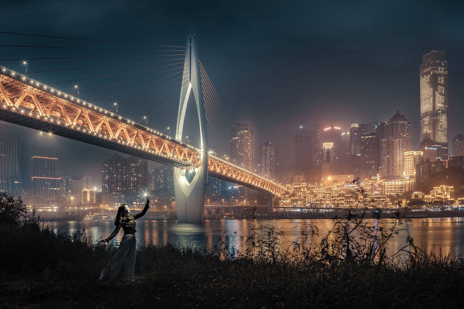 Chongqing 桌布 壁紙 — 夜晚，一個孤獨的身影站在河岸上，在發光的斜張橋下舉起雙臂，背後是充滿活力的城市天際線倒映在水中。