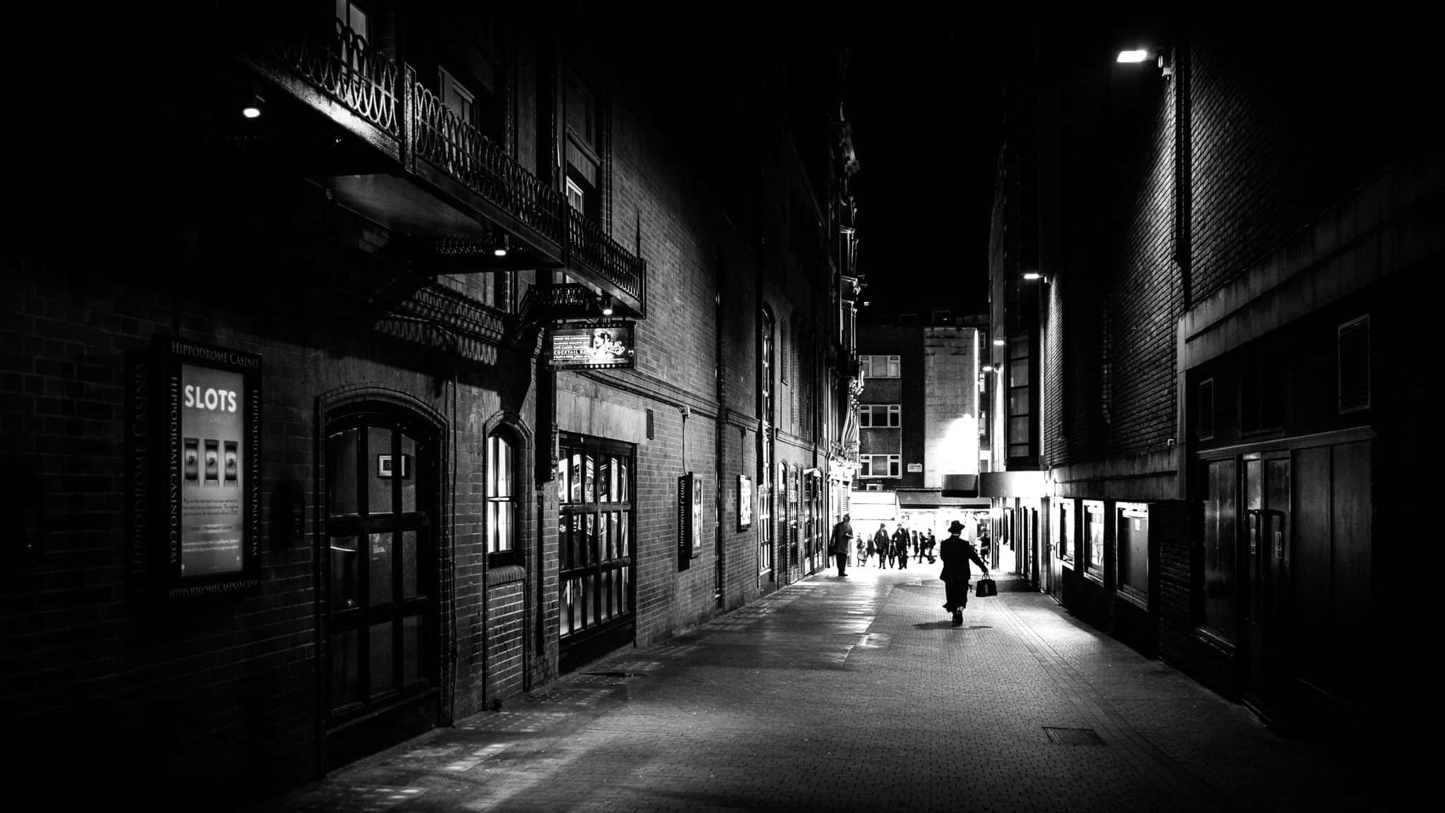 Black And White Street 桌布 壁紙 — 夜晚，一個孤獨的身影走在一條狹窄、燈光昏暗的城市小巷中，磚牆和散落的街燈投下長長的影子。
