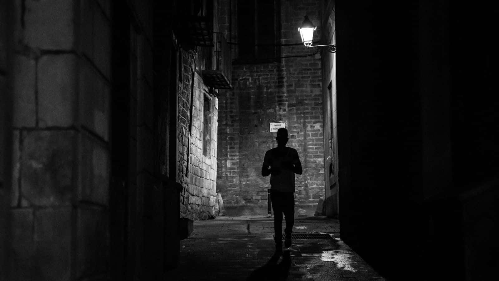 Dark Alleyway 桌布 壁紙 — 夜晚，一個孤獨的身影走在一條狹窄的石巷中，由一盞懸掛的燈籠照亮，投下長長的影子。