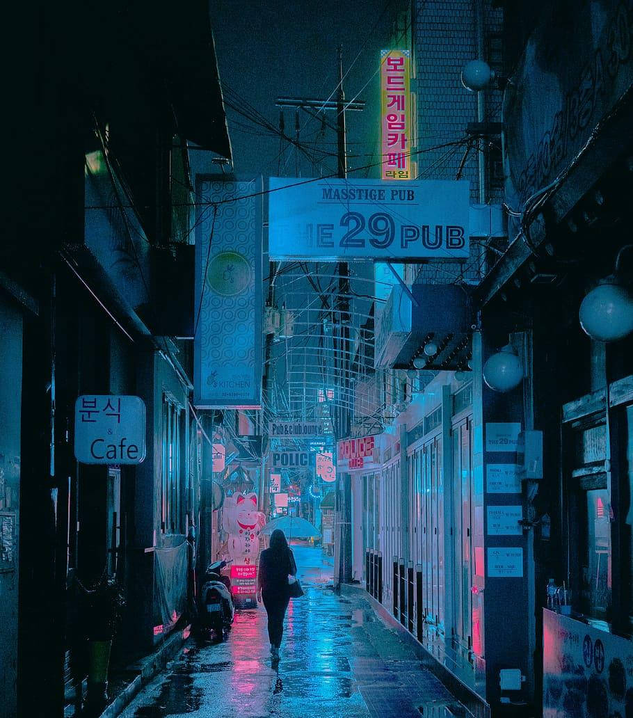 korean 桌布 壁紙 — 夜晚，一個孤獨的身影走在一條霓虹燈照亮的小巷中，雨水濕滑的路面反射著韓國酒吧和咖啡館鮮豔的藍色和粉紅色招牌。
