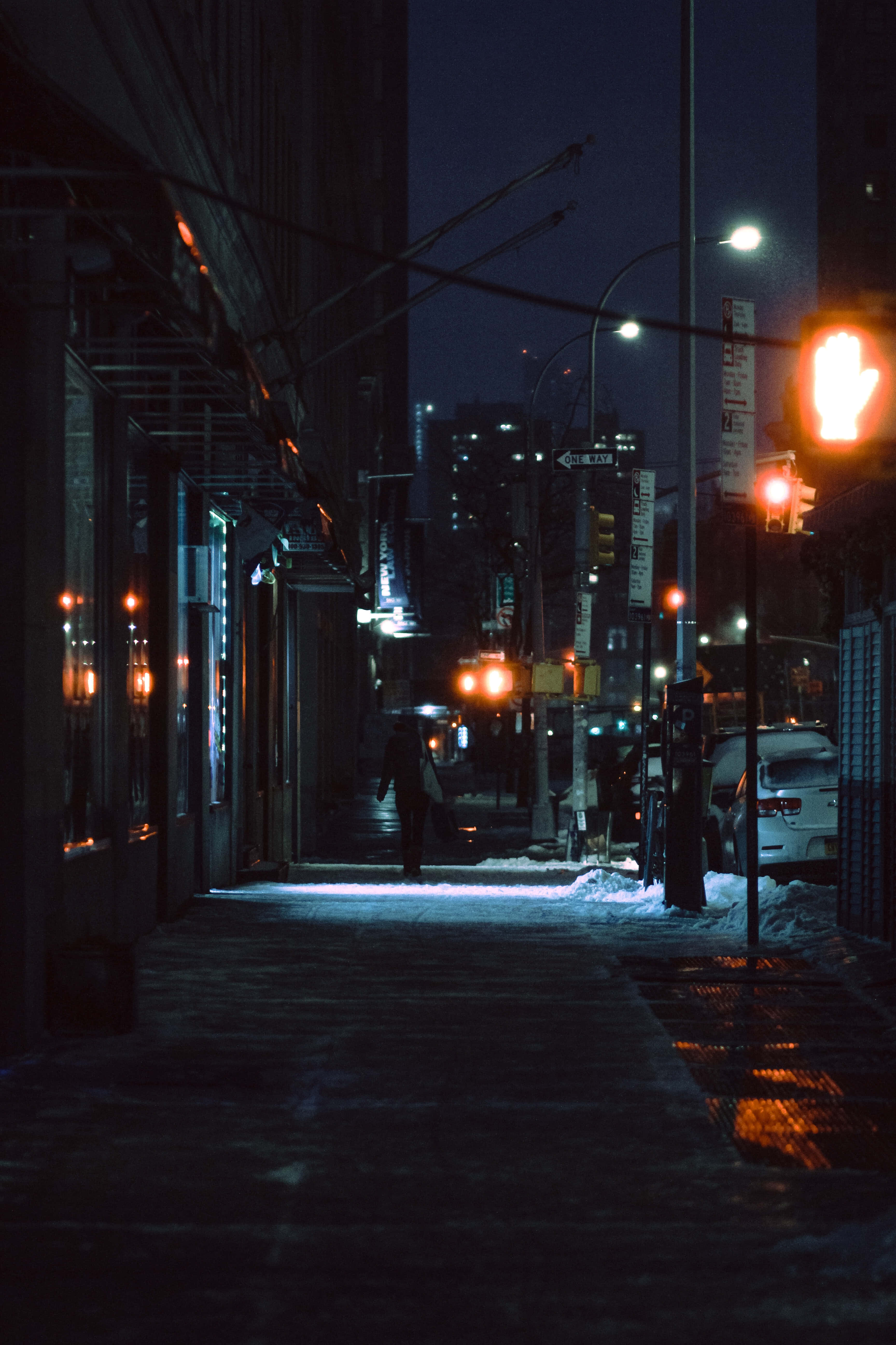 Night Time 桌布 壁紙 — 夜晚，一個孤獨的身影走在積雪的城市人行道上，被發光的紅色交通號誌和路燈照亮。