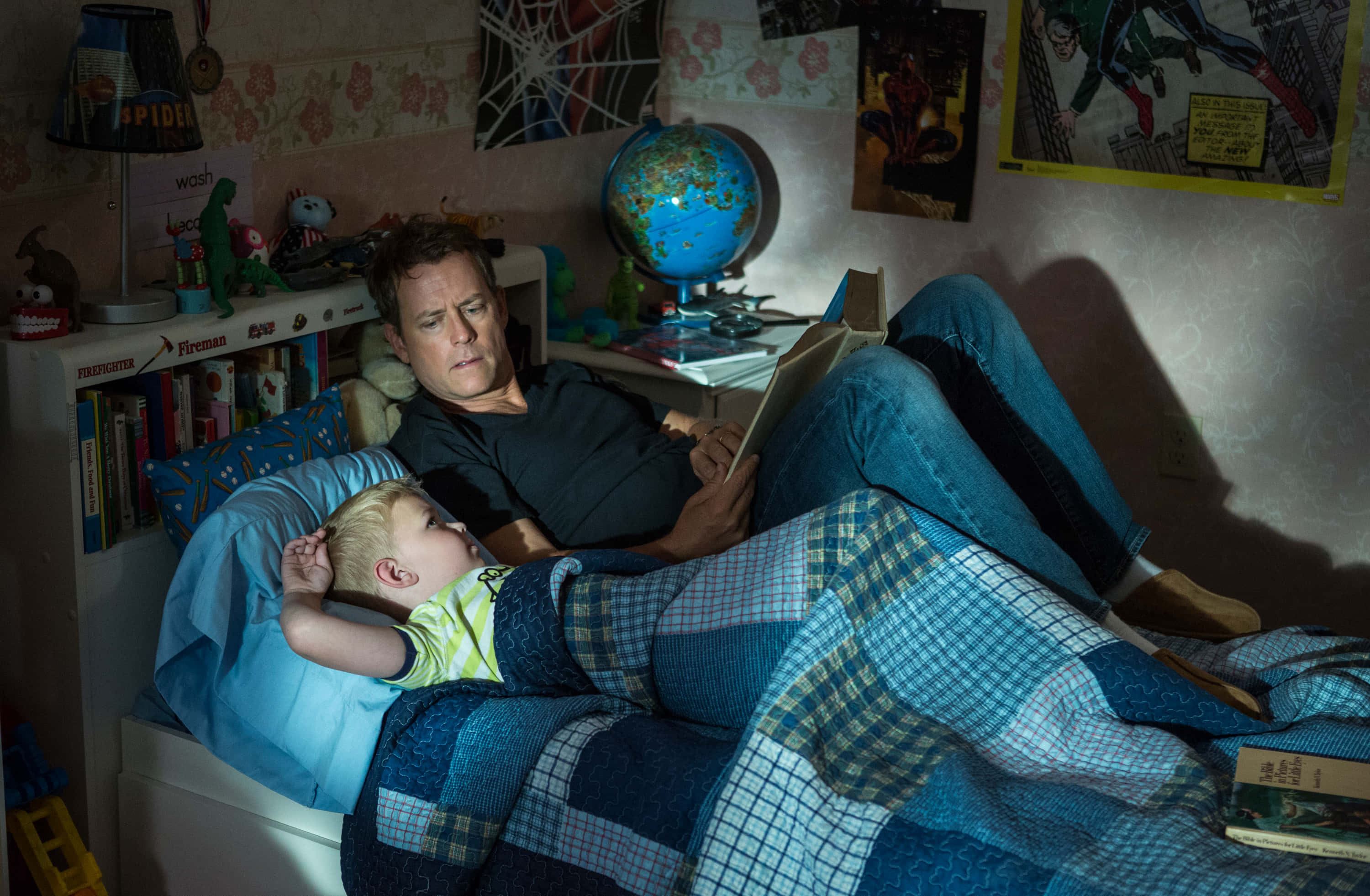 Greg Kinnear 桌布 壁紙 — 夜晚，一個男人在舒適雜亂的兒童臥室裡，在藍色被子下給一個孩子讀書。