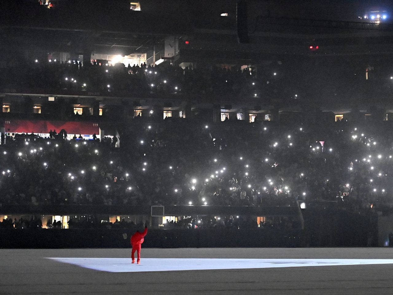 Kanye West Donda 桌布 壁紙 — 夜晚，一個身穿紅色衣服的孤獨身影站在舞台中央，在明亮的燈光下，體育場的人群舉起發光的手機螢幕。
