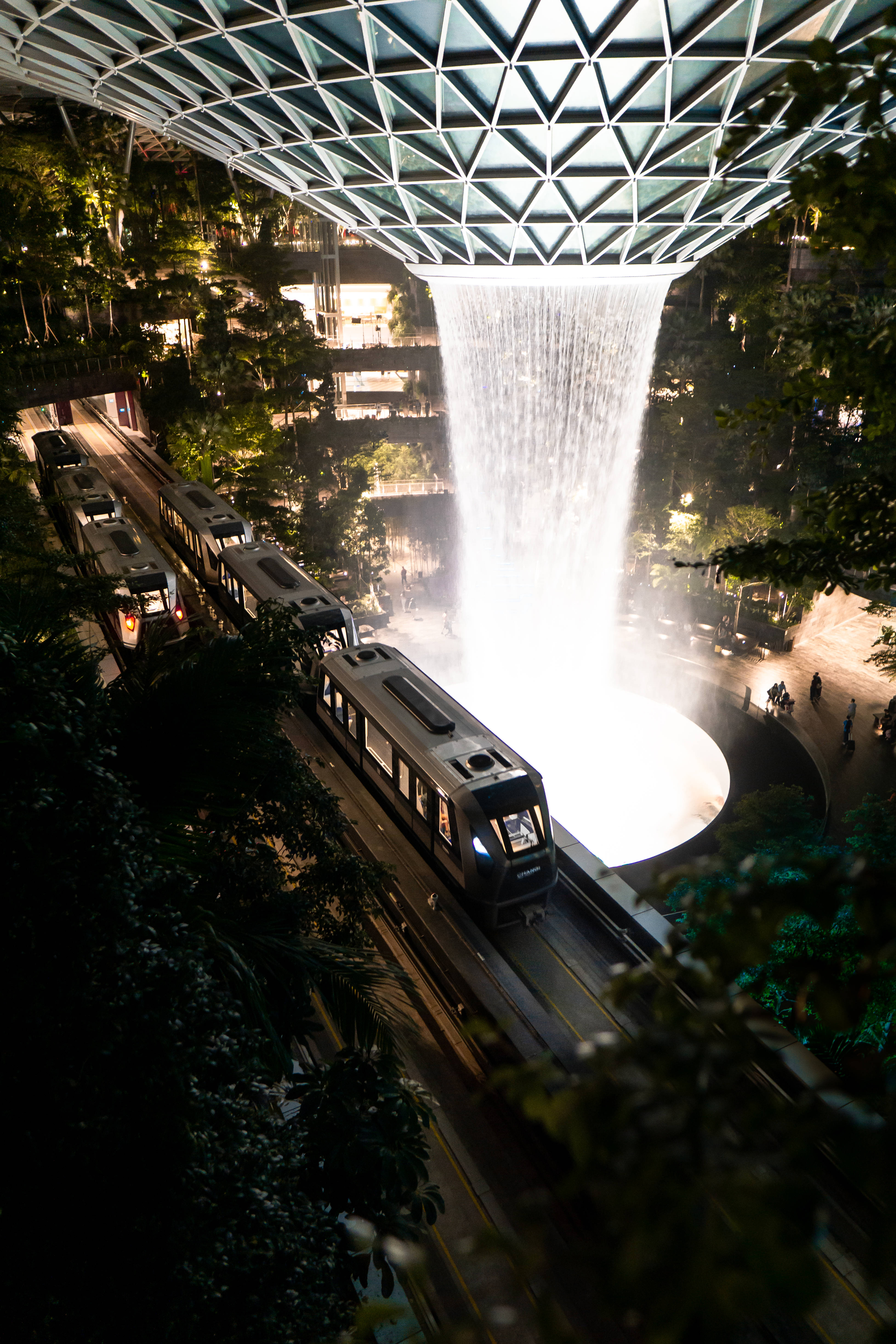 藝術 iPhone 桌布 壁紙 — 夜晚，一列火車在幾何玻璃天花板下，於一個大型室內瀑布下方滑行而過。