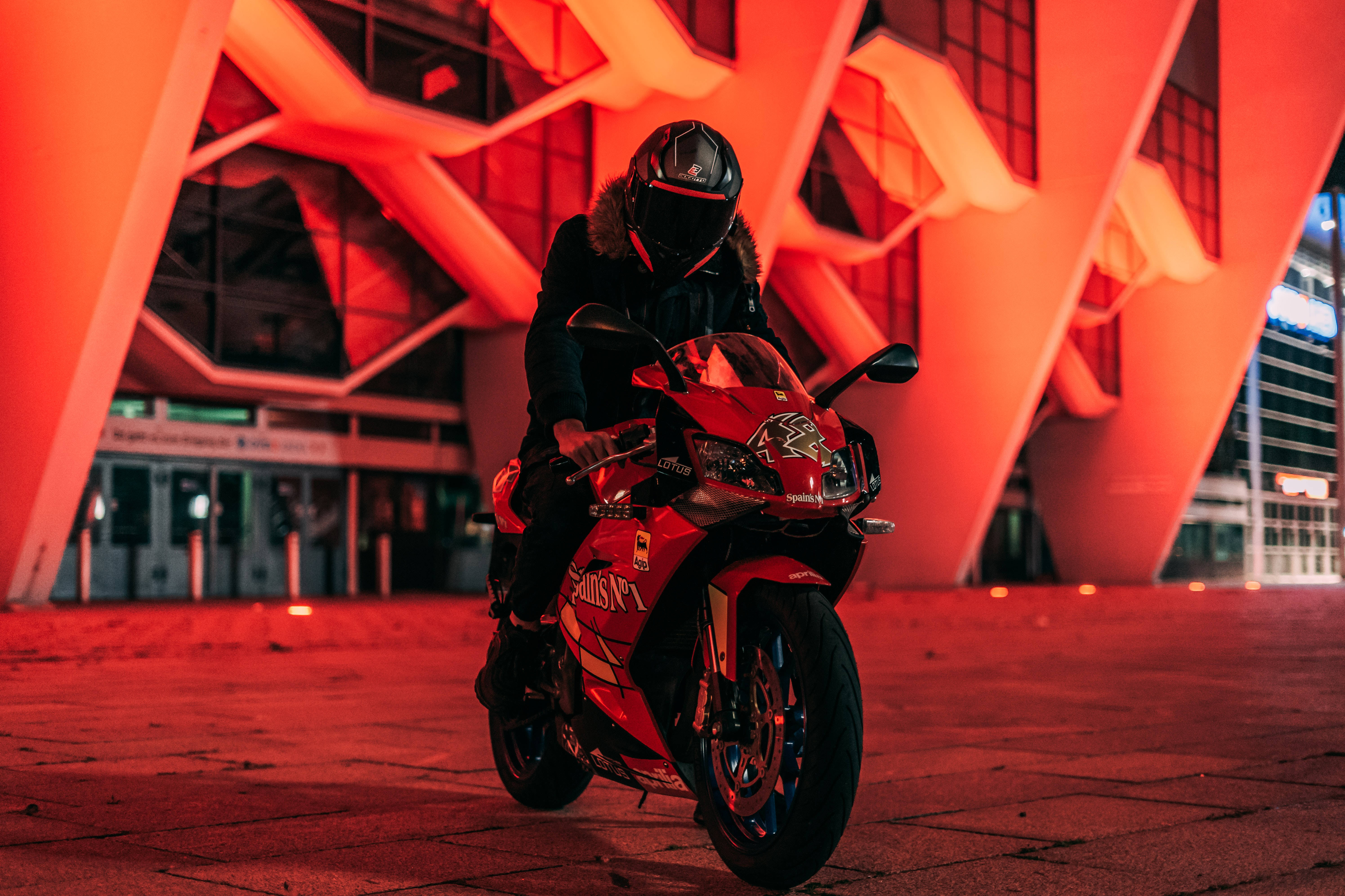 機車 桌布 壁紙 — 夜晚，一名騎士在紅色運動機車上，在發光的橙色幾何建築前暫停。
