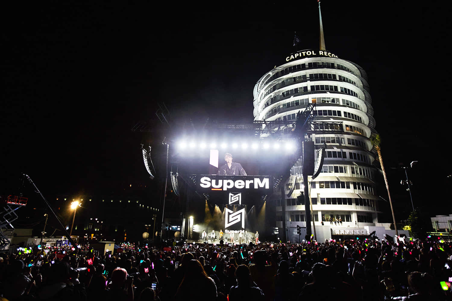 Party 桌布 壁紙 — 夜晚，一大群人在明亮的舞台燈光下聚集參加SuperM演唱會，標誌性的Capitol Records大樓在背後閃耀。