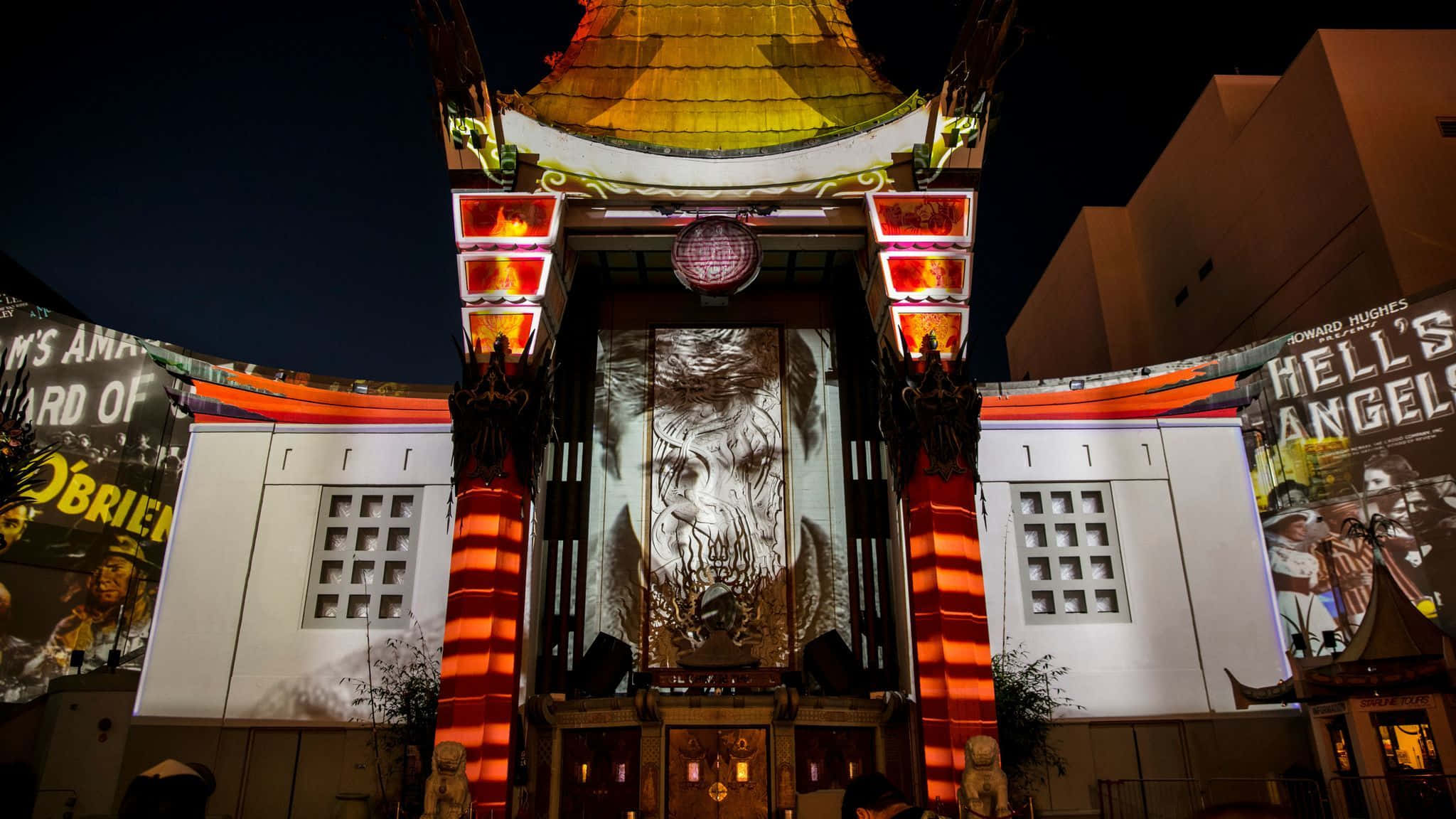 電影動圖 桌布 壁紙 — 夜晚，一座宏偉的劇院外牆被火紅燈光照亮，中央螢幕上投射出一個巨大的怪物面孔。