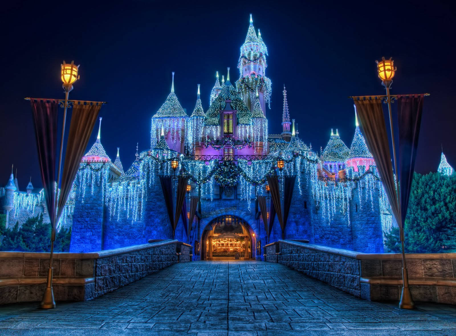 Disney Castle 桌布 壁紙 — 夜晚，一座宏偉的城堡被藍色和紫色的燈光照亮，從發光燈籠下的石橋上觀看。