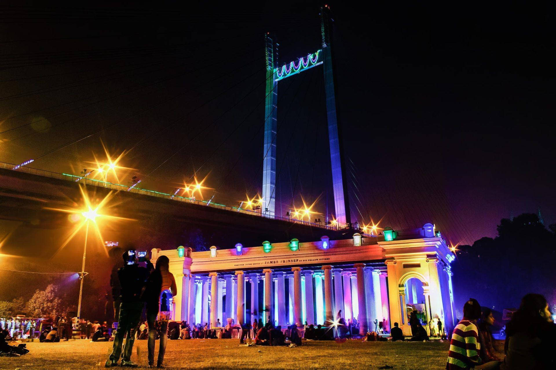 Kolkata 桌布 壁紙 — 夜晚，一座宏偉的燈光橋樑聳立於古典建築之上，人群在充滿活力的燈光下聚集在草地上。