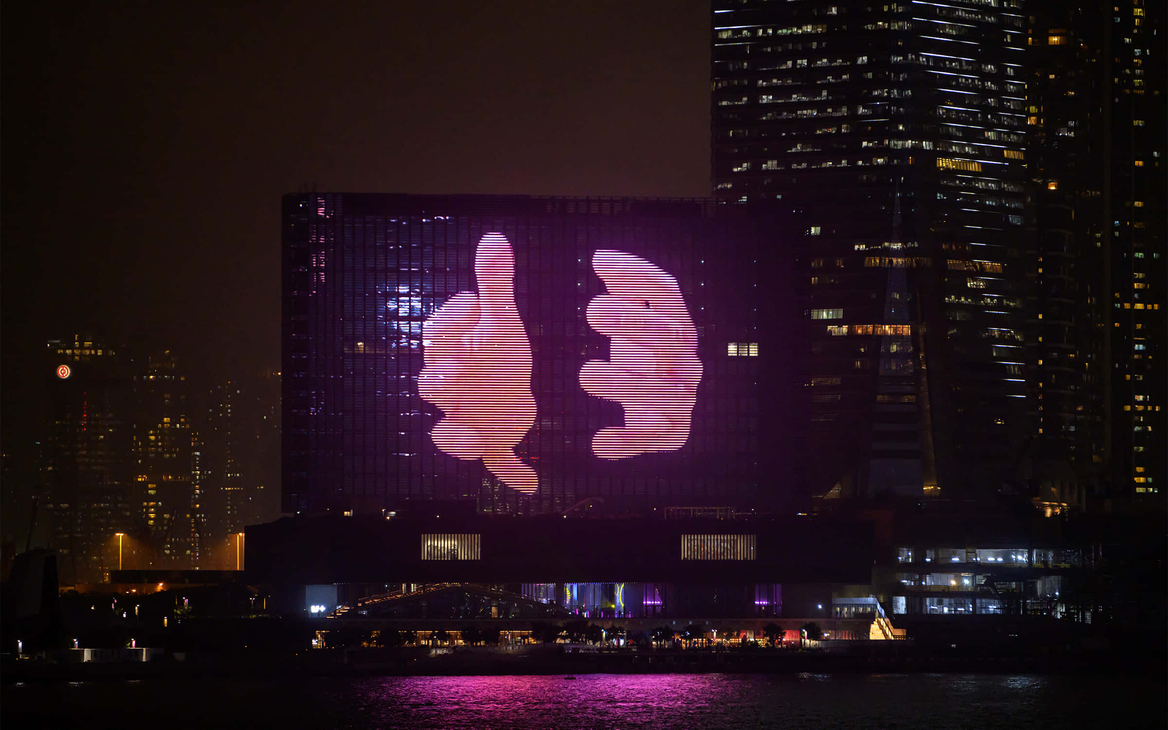 香港 桌布 壁紙 — 夜晚，一座巨大建築物的外牆在 illuminated 摩天大樓和黑暗水面倒影的背景下，顯示出一個發光的粉紅色豎起大拇指和放下大拇指的手勢。