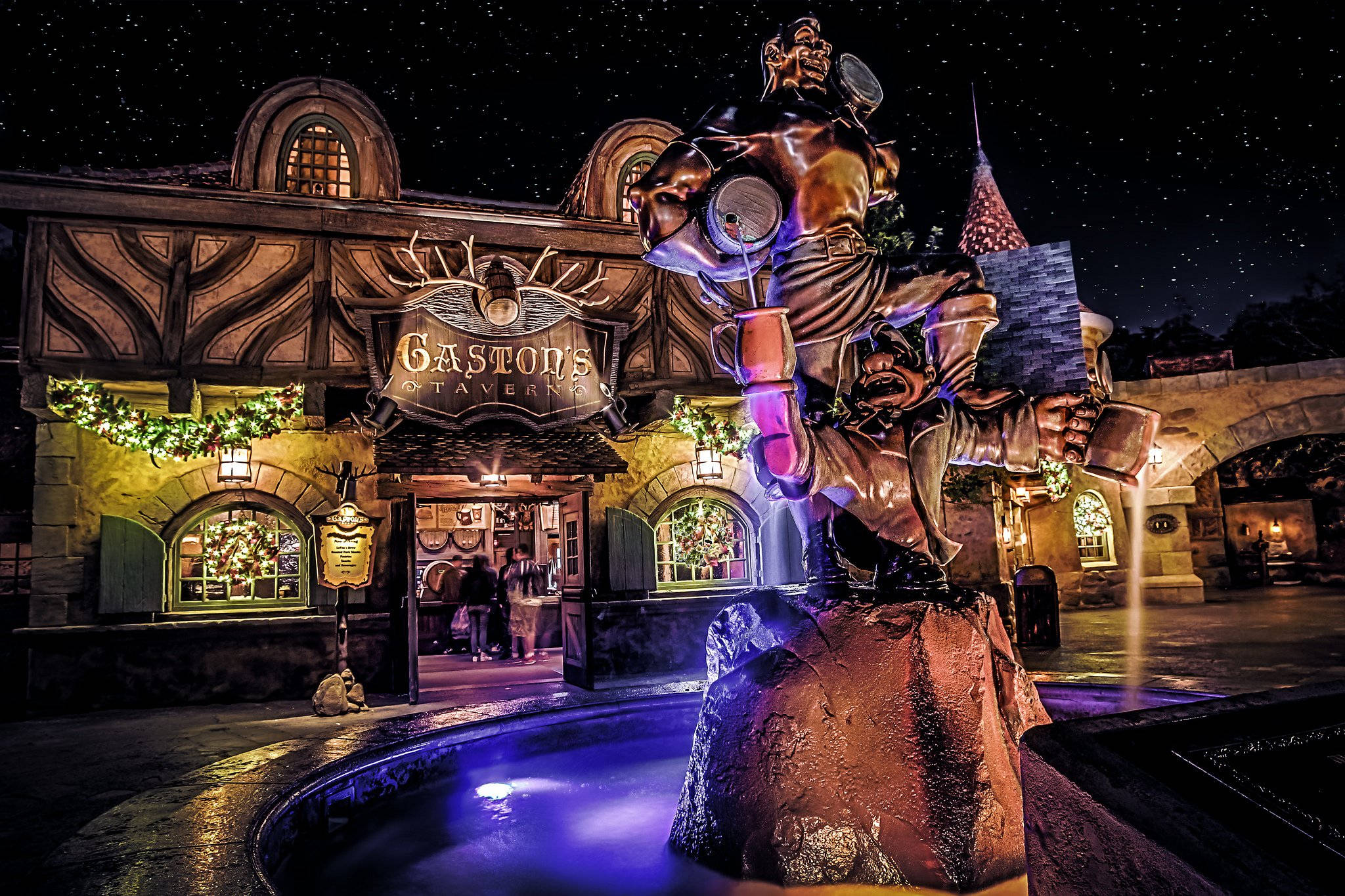 Universal 桌布 壁紙 — 夜晚，一座巨大的加斯頓銅像矗立在噴泉頂上，被紫色燈光照亮，襯托著滿天繁星，後方的加斯頓酒館散發著溫暖的光芒。