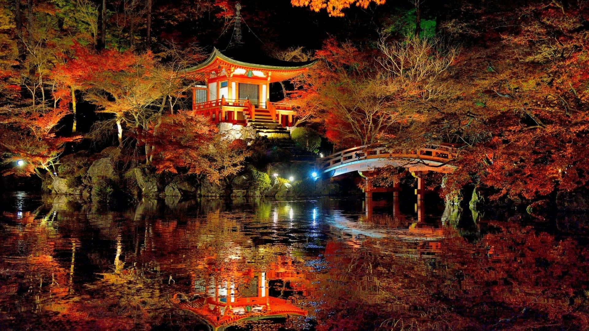日本 桌布 壁紙 — 夜晚，一座發光的紅色日本寺廟和拱橋倒映在平靜的水面上，周圍是火紅的秋葉。
