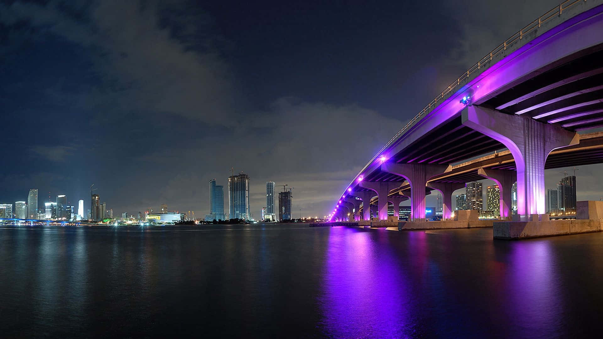 Miami 桌布 壁紙 — 夜晚，一座發光的紫色橋樑拱在平靜的水面上，城市天際線倒映在黑暗的水面。