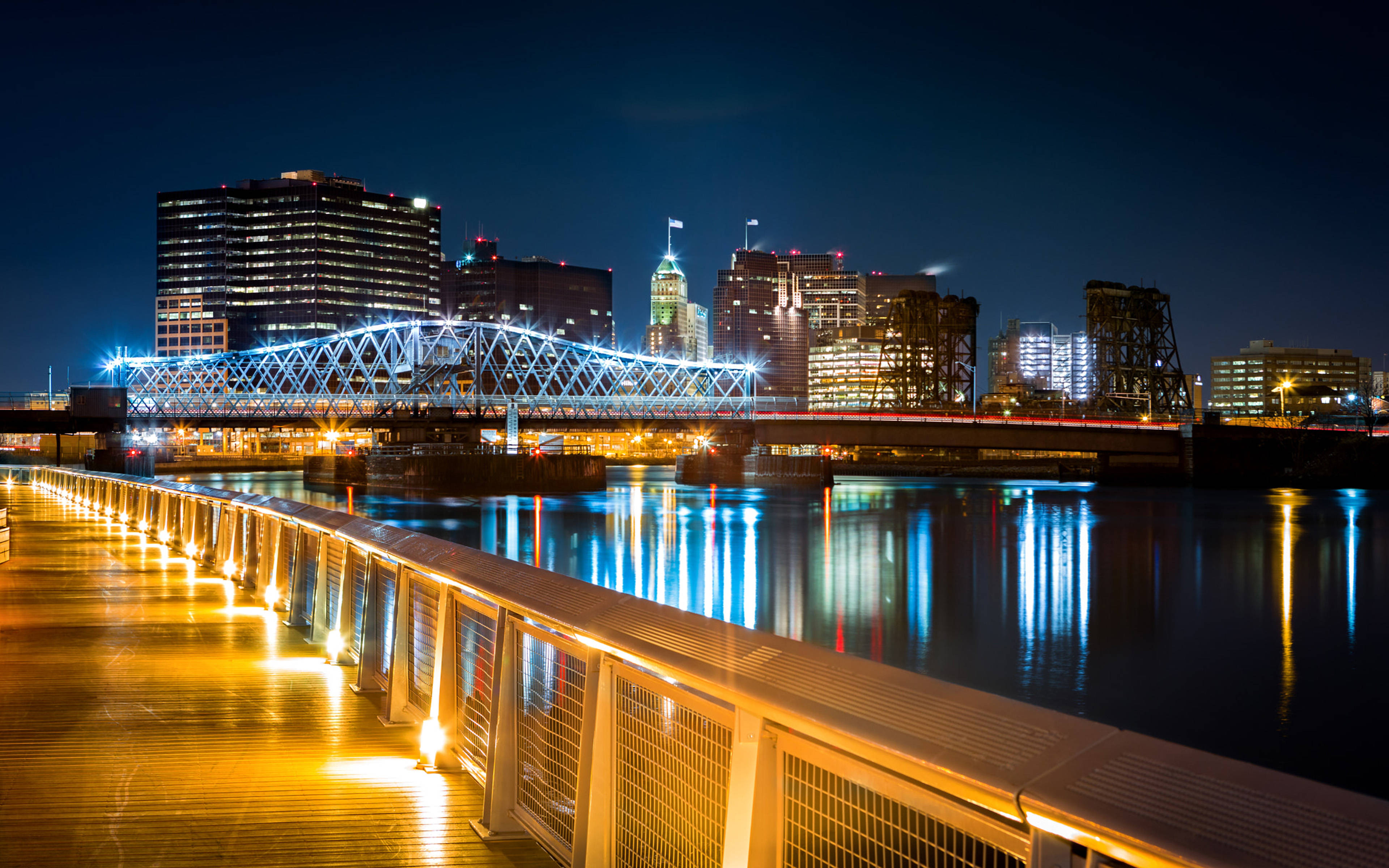 bridge 桌布 壁紙 — 夜晚，一座發光的藍色桁架橋橫跨河流，城市燈光倒映在水中，前景是溫暖照亮的步道。
