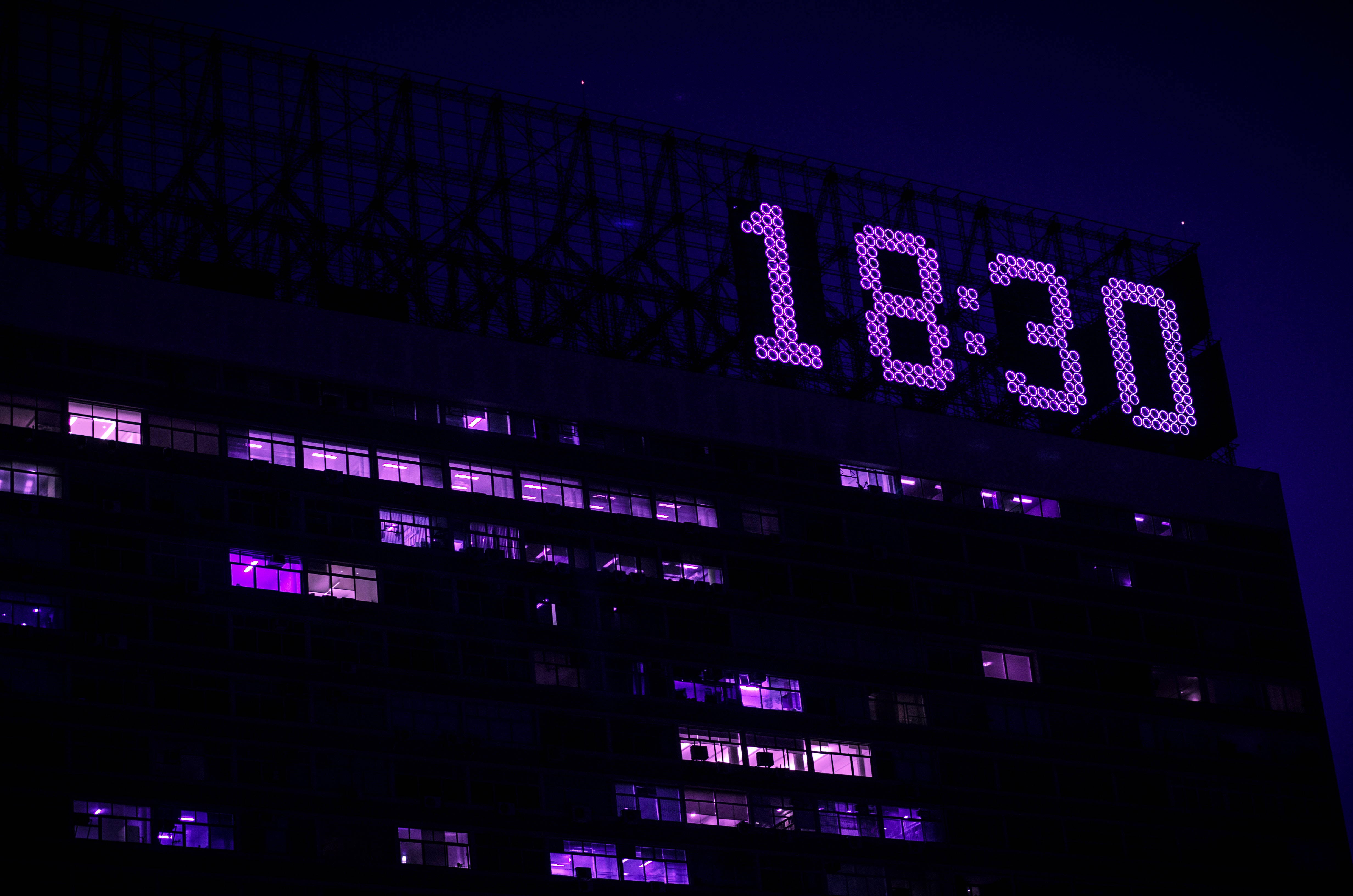 霓虹紫 iPhone 桌布 壁紙 — 夜晚，一座高聳的建築物發光，上面有一個巨大的紫色數字時鐘，在明亮的窗戶上方顯示18:30。