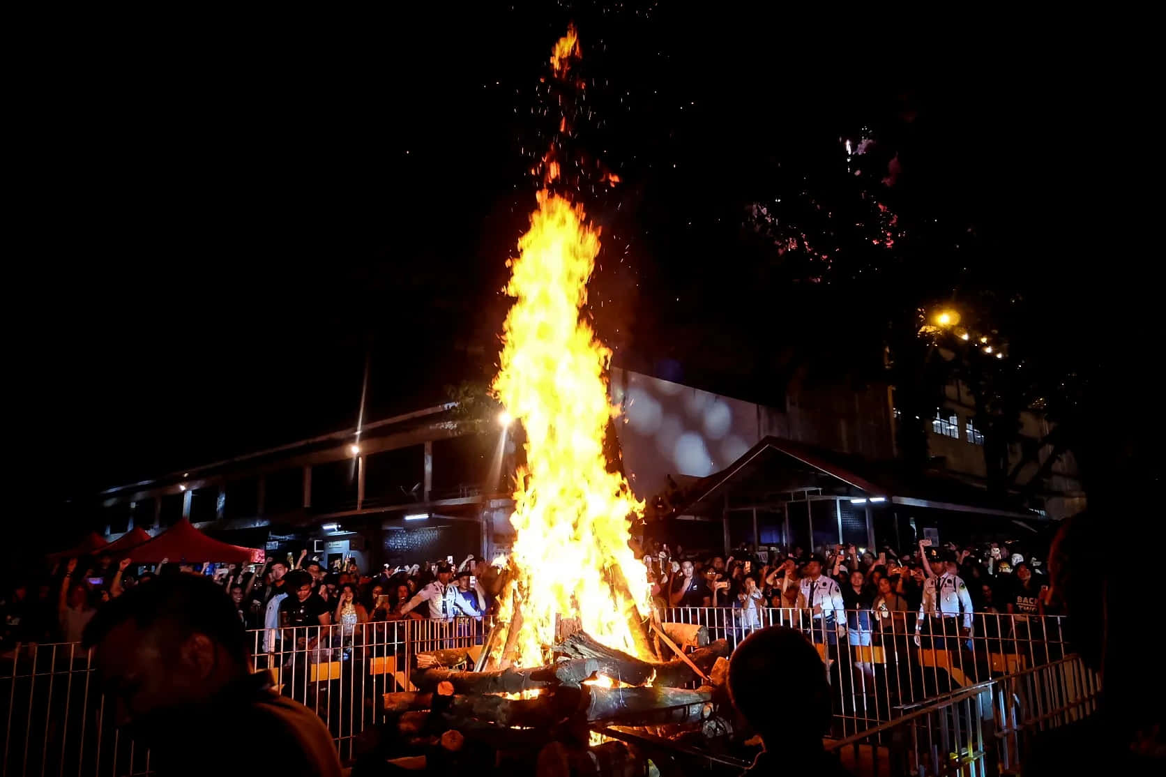 Bonfire 桌布 壁紙 — 夜晚，一座高聳的營火噴發出明亮的橙色火焰，照亮了聚集在圍欄後的人群。