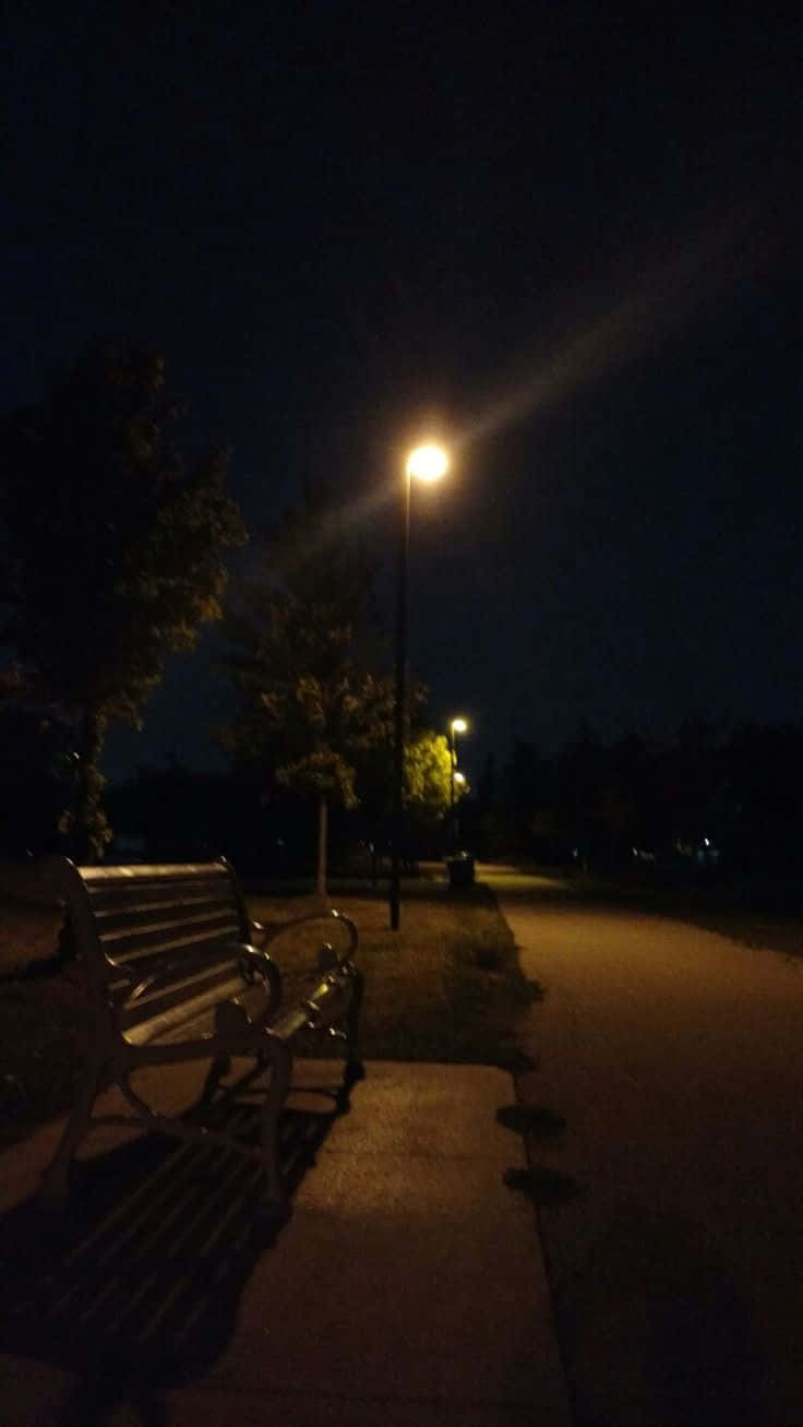 Night Time 桌布 壁紙 — 夜晚，一張孤獨的公園長椅坐在發光的街燈下，在寧靜的小徑上投下長長的影子。