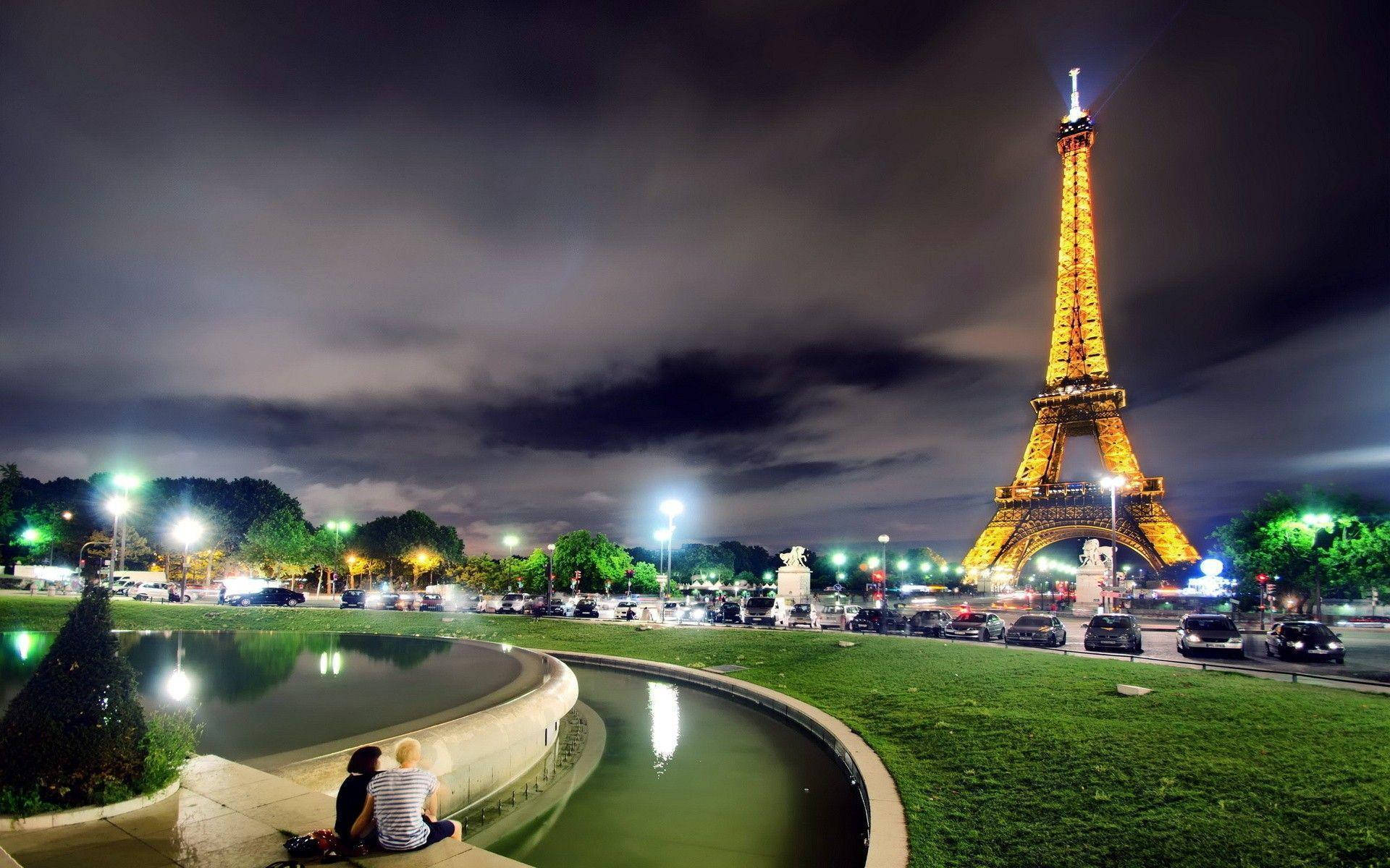 Eiffel Tower 桌布 壁紙 — 夜晚，兩個人坐在彎曲的噴泉旁，艾菲爾鐵塔在陰雲密佈的天空下閃耀著金色光芒。