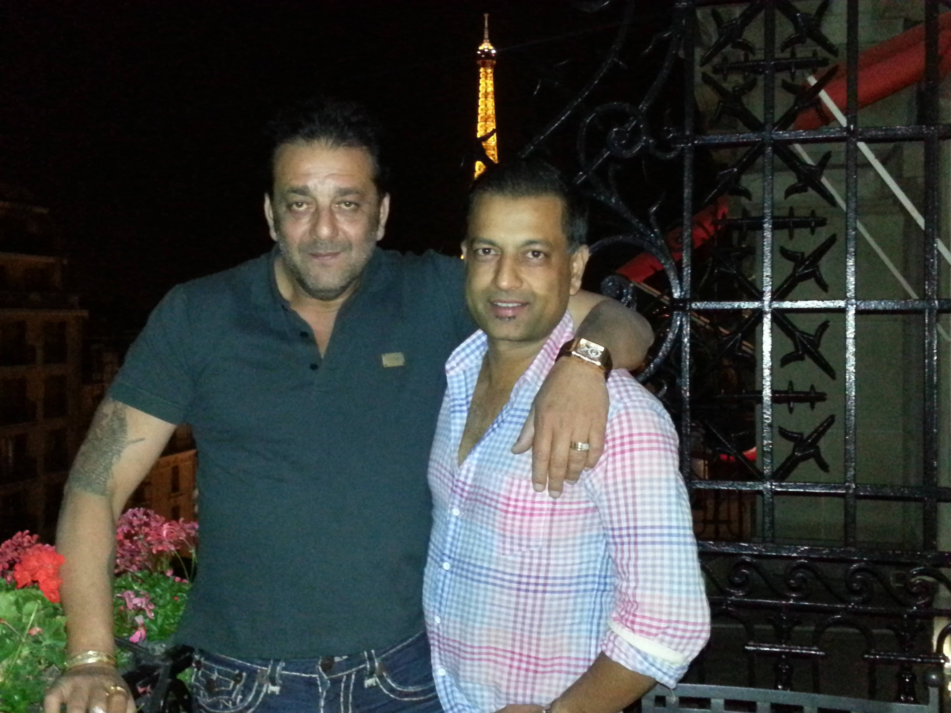 Sanjay 桌布 壁紙 — 夜晚，兩個男人在裝飾華麗的黑色大門後面，以閃爍的艾菲爾鐵塔為背景拍照。