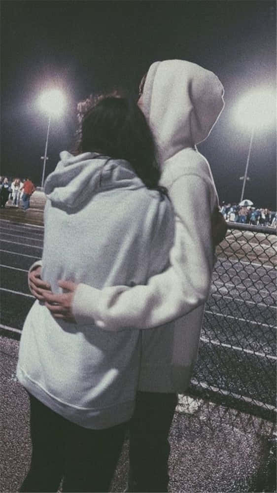 Girlfriend And Boyfriend 桌布 壁紙 — 夜晚，兩個穿著連帽衫的人在明亮的體育場燈光下，在鐵絲網圍欄附近溫暖地擁抱。
