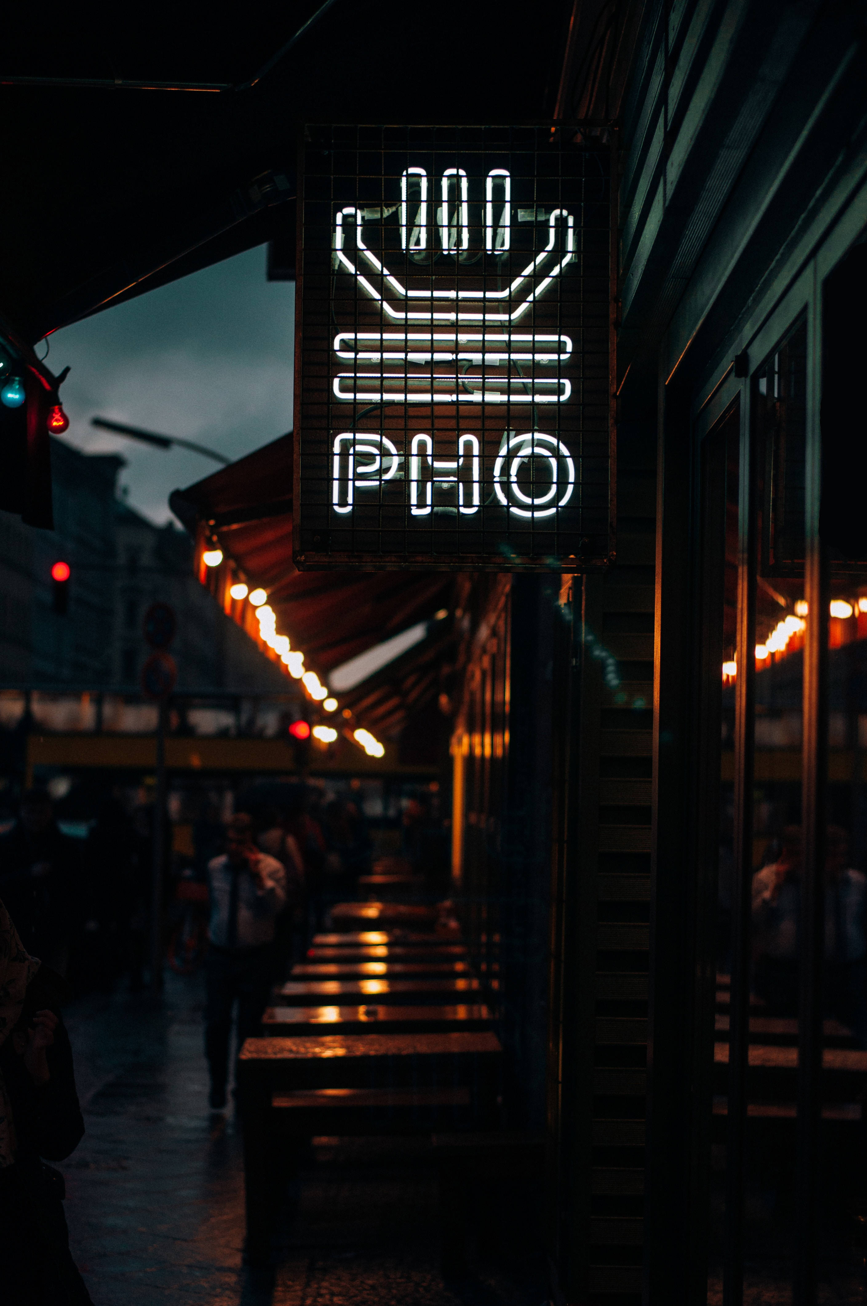 霓虹城市動態圖 桌布 壁紙 — 夜晚，在溫暖燈光和模糊行人背景下，一隻發光的手形霓虹燈牌懸掛在越南河粉餐廳上方。