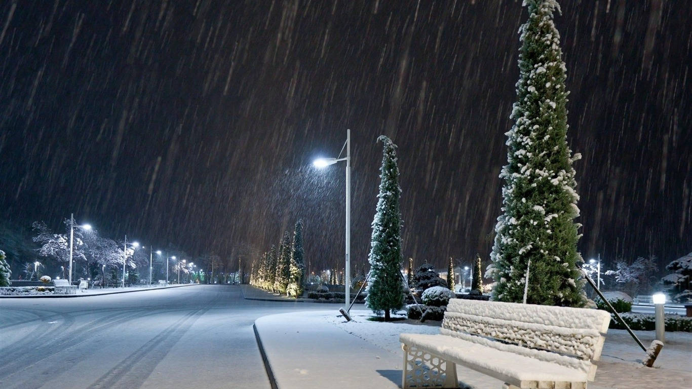 冬天 桌布 壁紙 — 夜晚，大雪紛飛，一條寧靜的街道兩旁是燈火通明的樹木和一條積雪的長椅。