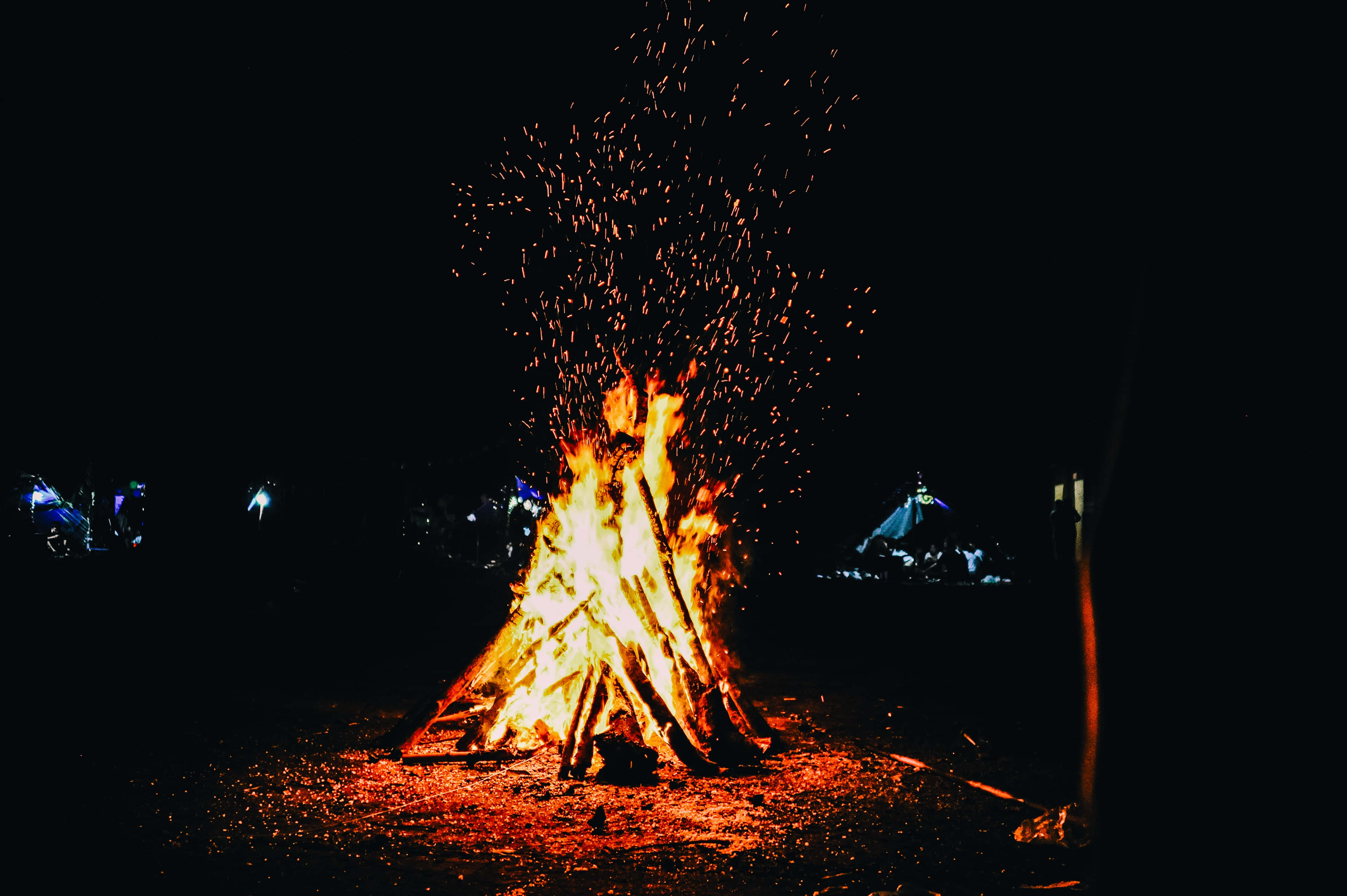Bonfire 桌布 壁紙 — 夜晚，熊熊燃燒的營火將明亮的橙色火花射向漆黑的天空，遠處的帳篷發出柔和的光芒。
