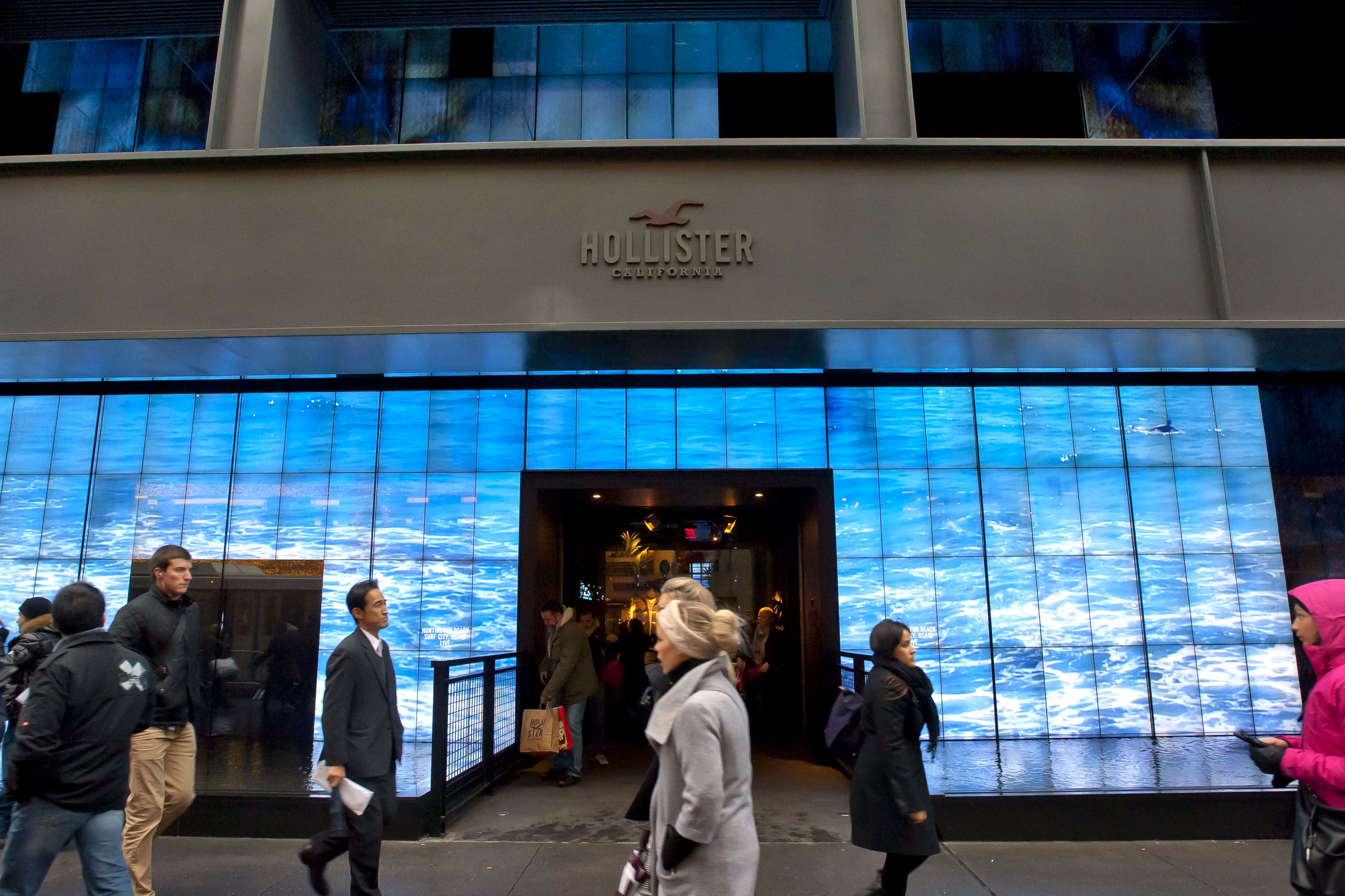 Hollister 桌布 壁紙 — 夜晚，行人經過一間Hollister商店，其外牆設有充滿活力的藍色海浪數位顯示屏。