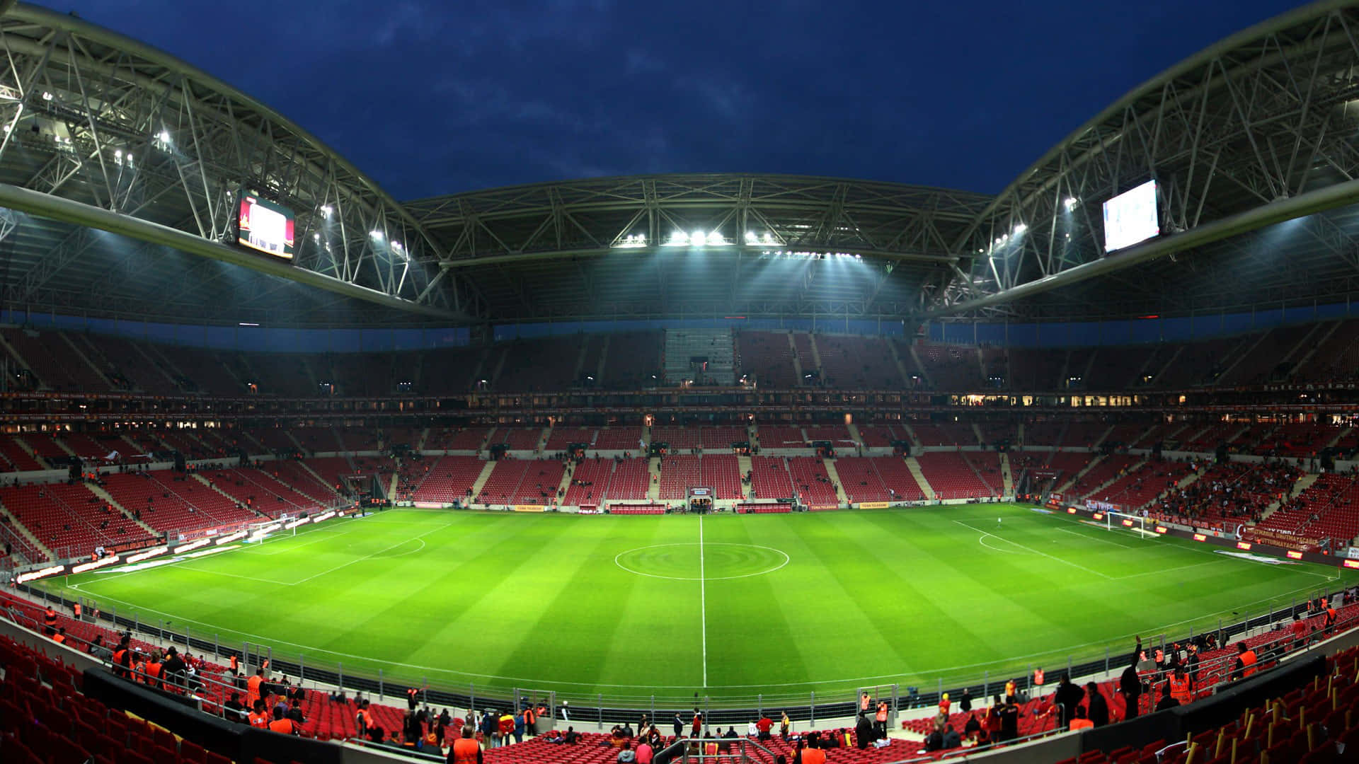 Stadium 桌布 壁紙 — 夜晚一個廣闊空蕩的足球場，明亮的泛光燈照亮了充滿活力的綠色球場和成排的紅色座位。