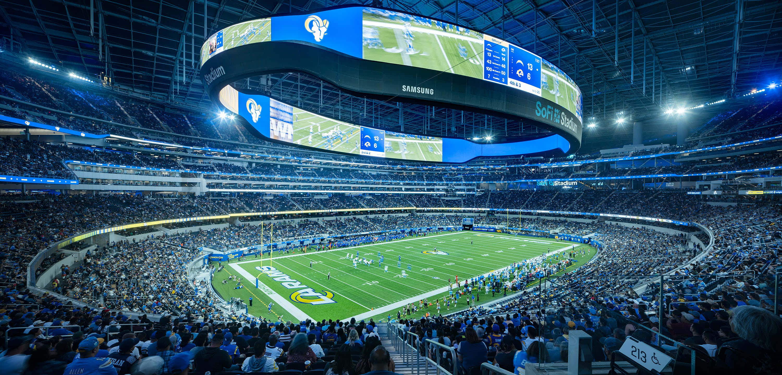Sofi Stadium 桌布 壁紙 — 夜晚一個擠滿人的足球場，明亮的藍色燈光照亮球場，上方有巨大的弧形螢幕。