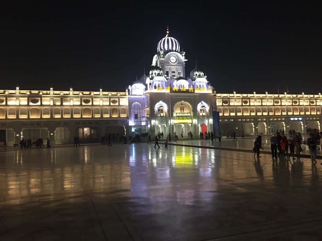 Golden Temple 桌布 壁紙 — 夜晚一座宏偉的錫克教謁師所，發光的圓頂和拱形窗戶倒映在濕漉漉的廣場上，人們在周圍漫步。