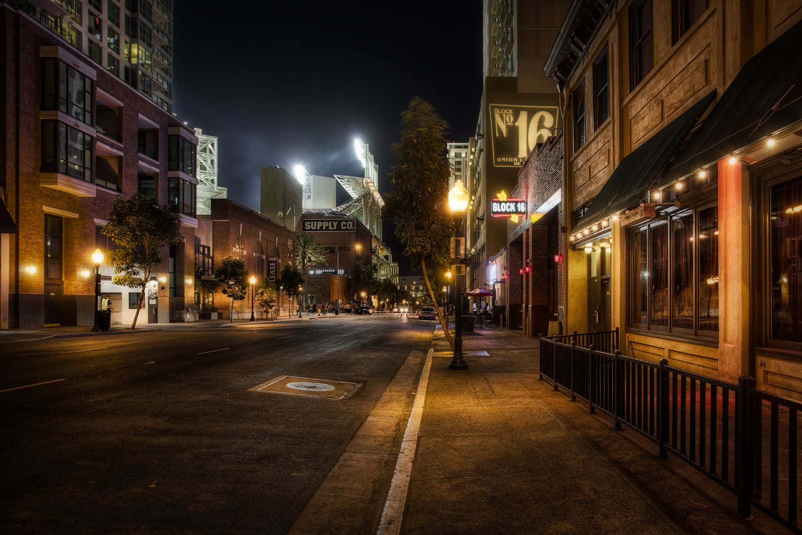 Night Street 桌布 壁紙 — 夜晚一條寧靜的城市街道，溫暖的路燈和發光的商店招牌在黑暗的天空下照亮。