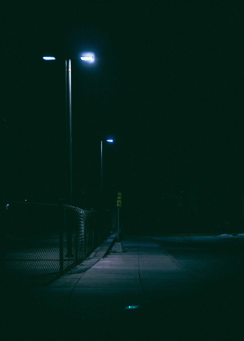 黑夜 桌布 壁紙 — 夜晚一條燈光昏暗的人行道，被冰冷的藍色街燈照亮，在鐵絲網圍欄上投下長長的影子。