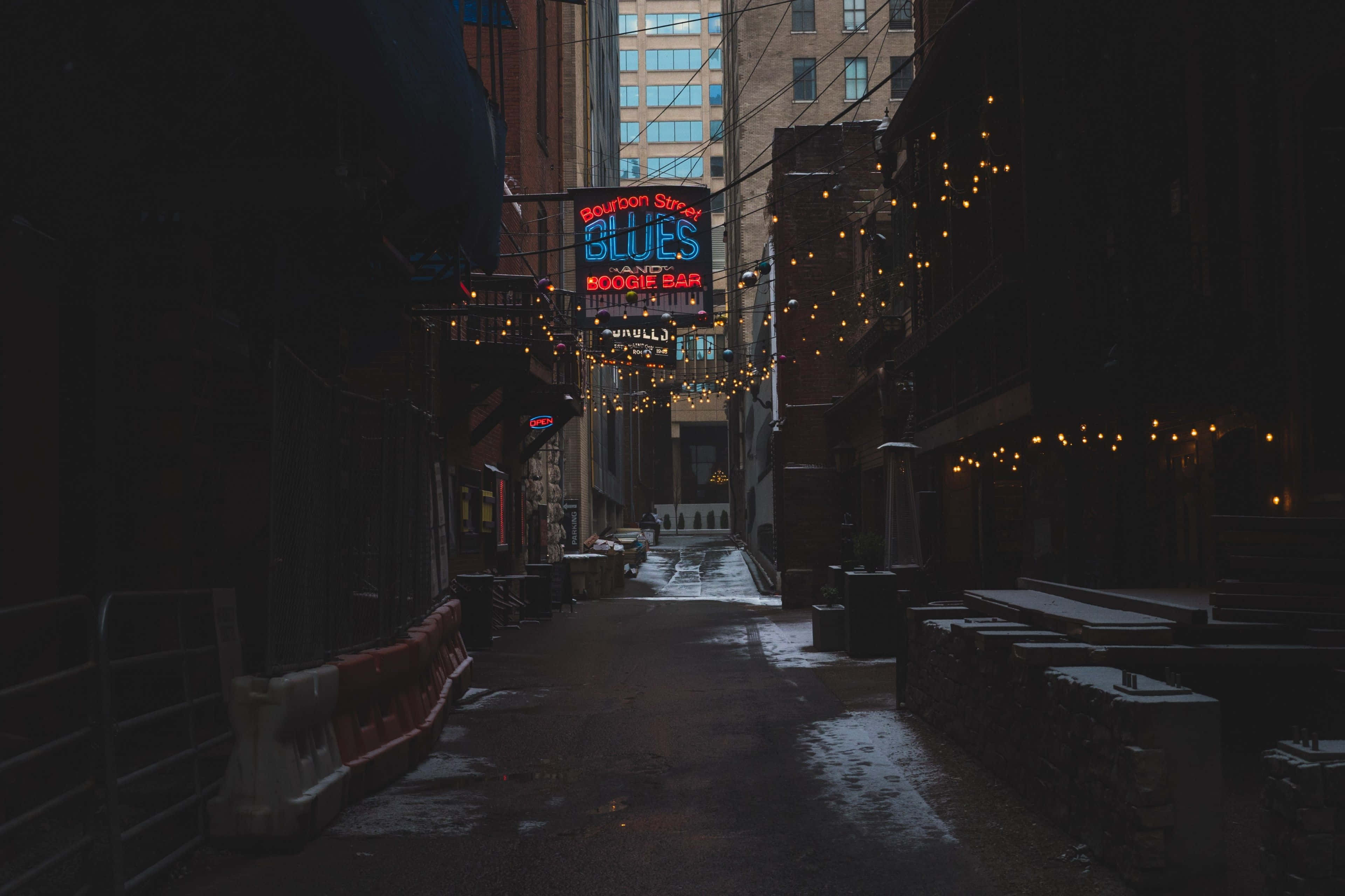 桌布 壁紙 — 夜晚一條燈光昏暗的城市小巷，有發光的 Bourbon Street Blues 霓虹招牌和串燈，在濕漉漉的路面上投下溫暖的光芒。