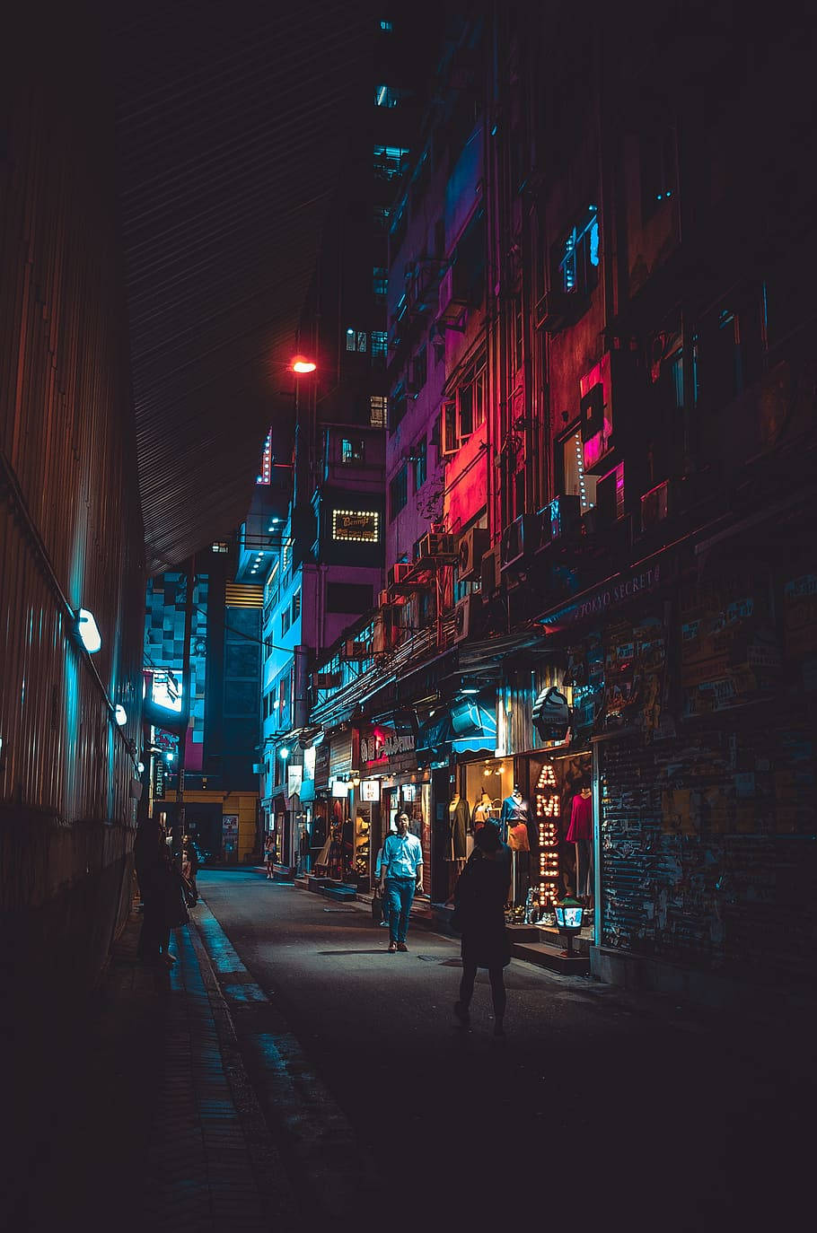 藝術 桌布 壁紙 — 夜晚一條狹窄小巷，被鮮豔的紅色和藍色霓虹燈招牌照亮，倒映在濕漉漉的人行道上，有行人走過。