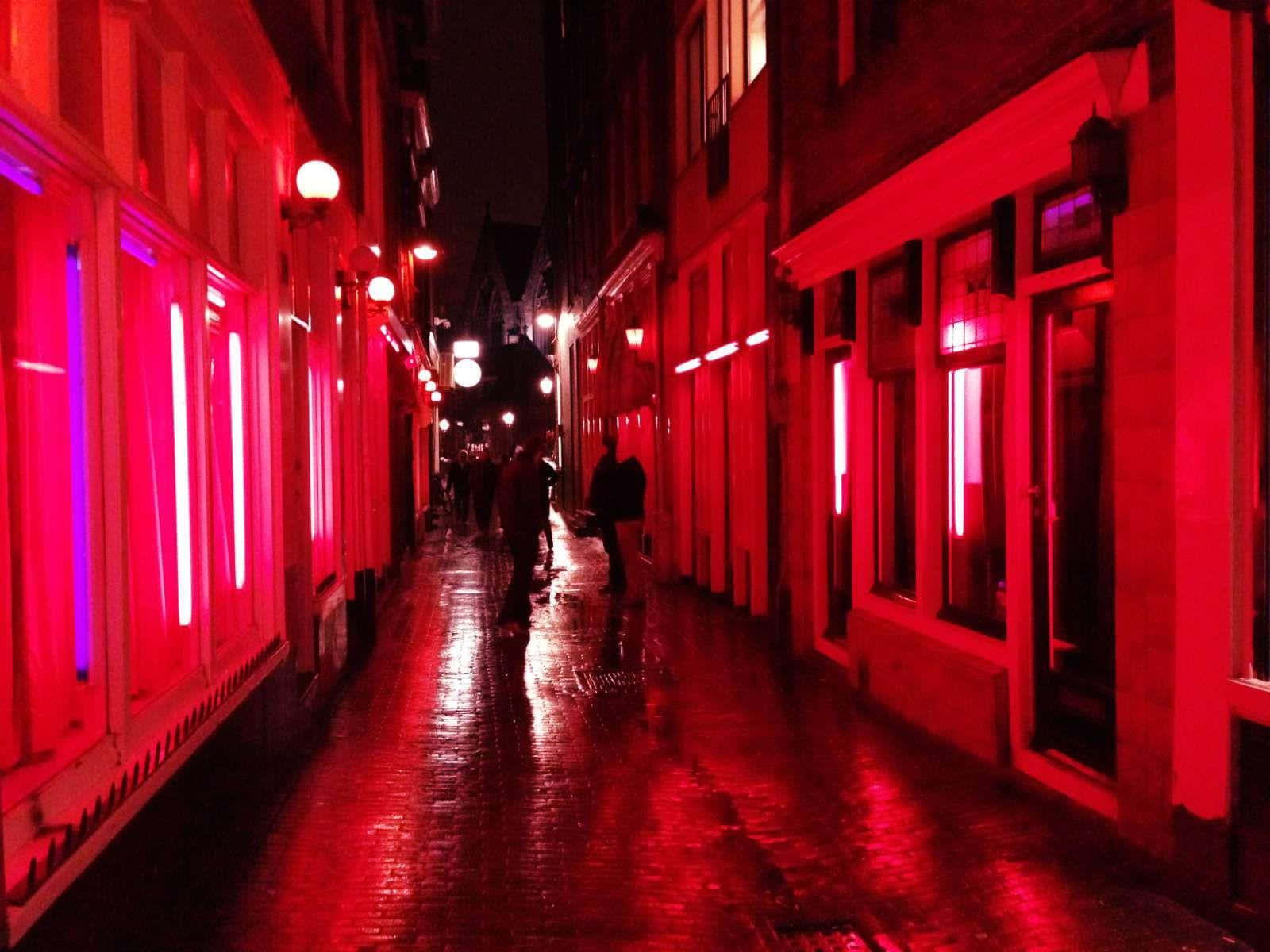 Red Light District 桌布 壁紙 — 夜晚一條狹窄潮濕的小巷，沐浴在強烈的紅色霓虹燈光中，燈光反射在鵝卵石上，行人形成剪影。