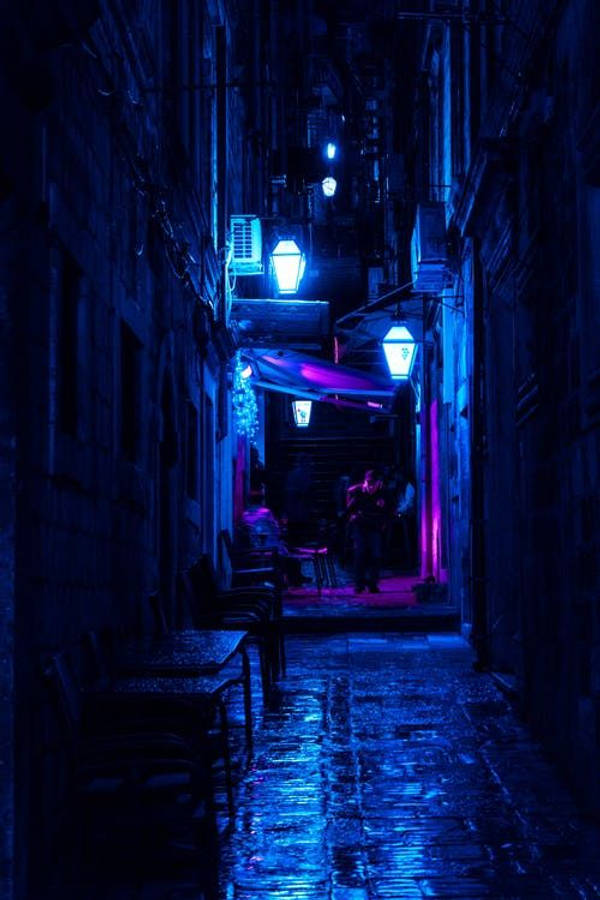 dark blue aesthetic tumblr 桌布 壁紙 — 夜晚一條狹窄潮濕的小巷，沐浴在電光藍色霓虹燈中，燈光反射在鵝卵石上，有剪影人物。