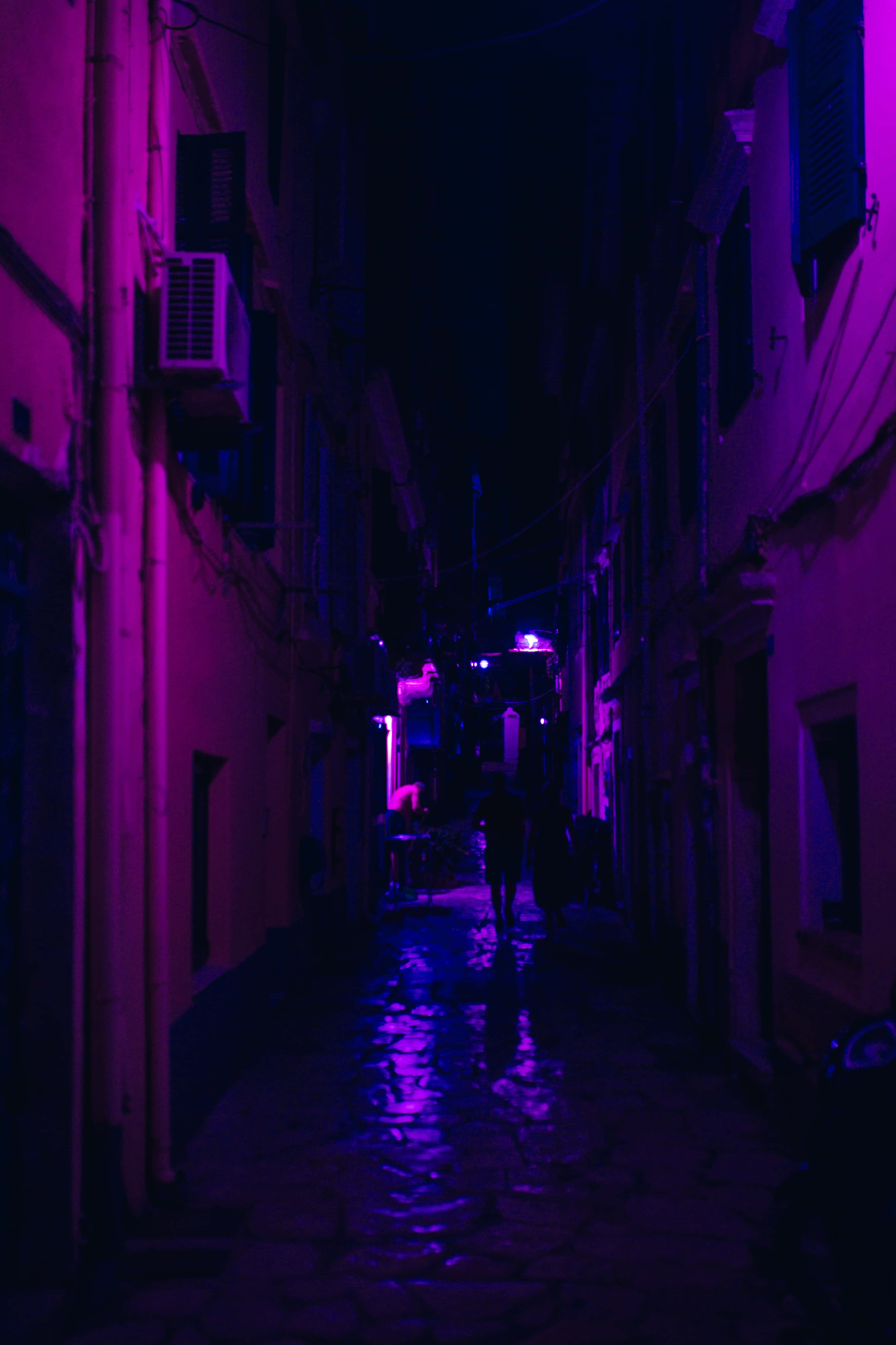 Dark Alleyway 桌布 壁紙 — 夜晚一條狹窄潮濕的小巷，沐浴在鮮豔的紫色霓虹燈光中，燈光在石路上反射，有剪影人物走遠。