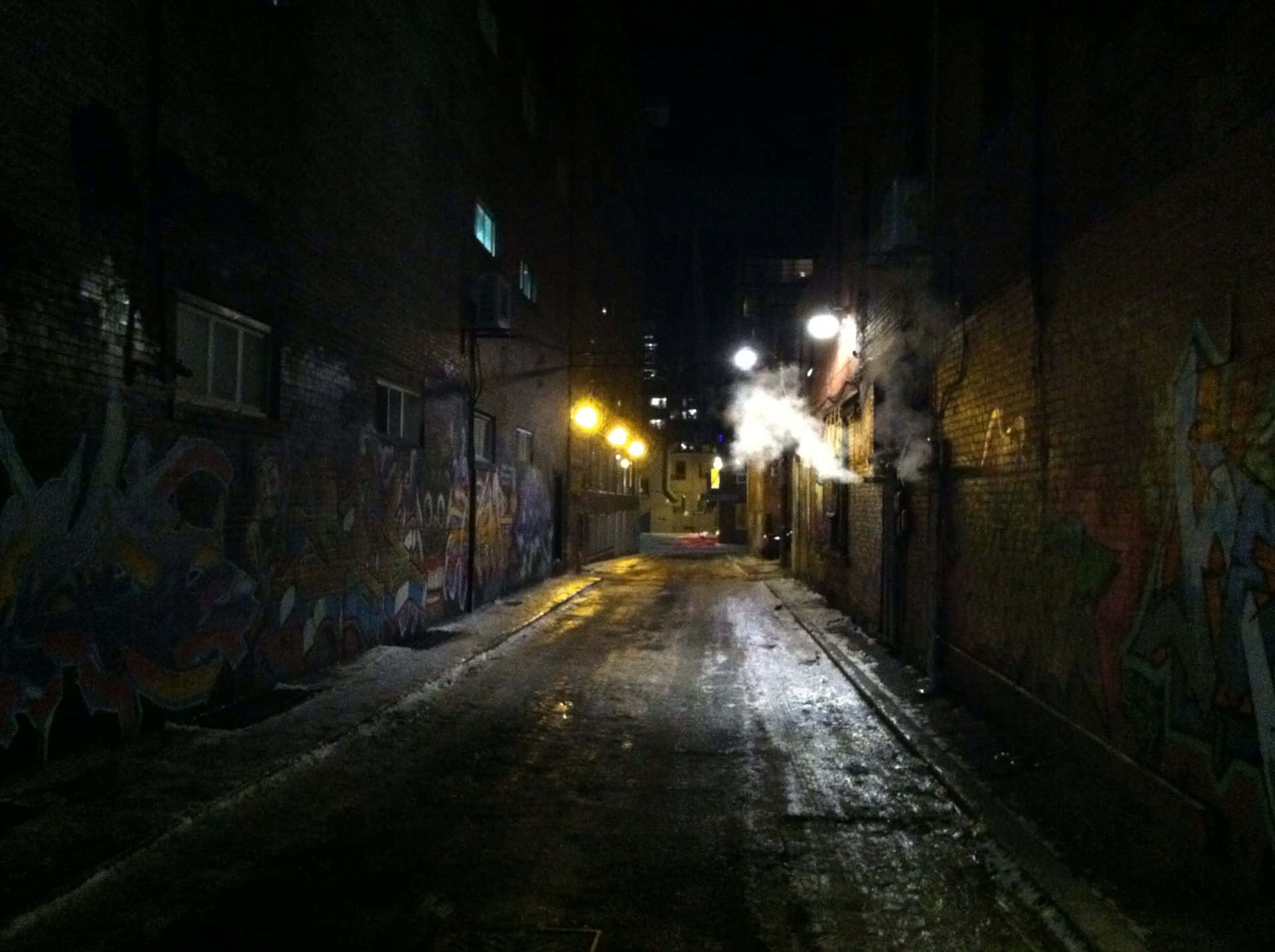Alleyway 桌布 壁紙 — 夜晚一條狹窄潮濕的小巷，街燈照亮，磚牆上有色彩繽紛的塗鴉，通風口升起蒸氣。