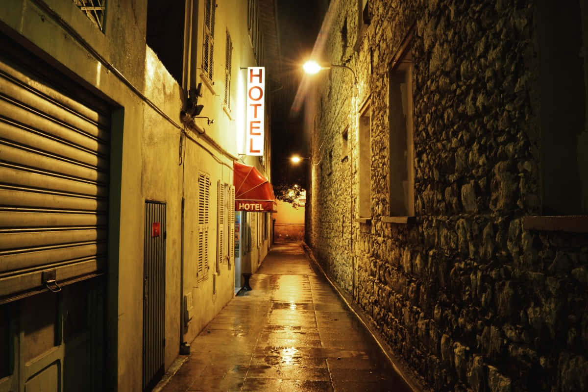 Alleyway 桌布 壁紙 — 夜晚一條狹窄潮濕的小巷，被發光的飯店招牌和路燈照亮，倒映在潮濕的鵝卵石上。