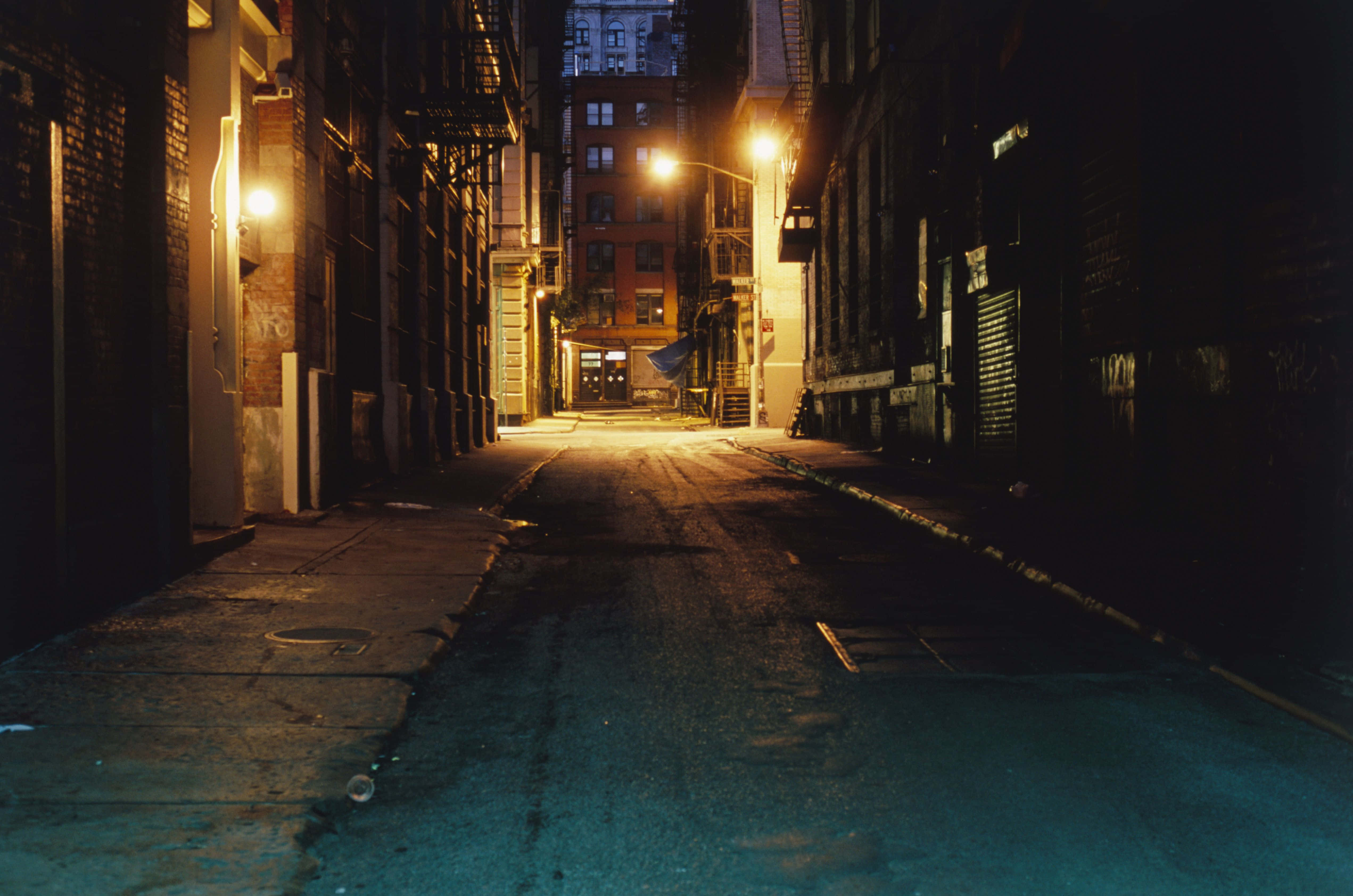 Alleyway 桌布 壁紙 — 夜晚一條狹窄的城市小巷，被溫暖的路燈照亮，在潮濕的路面上投下長長的影子。
