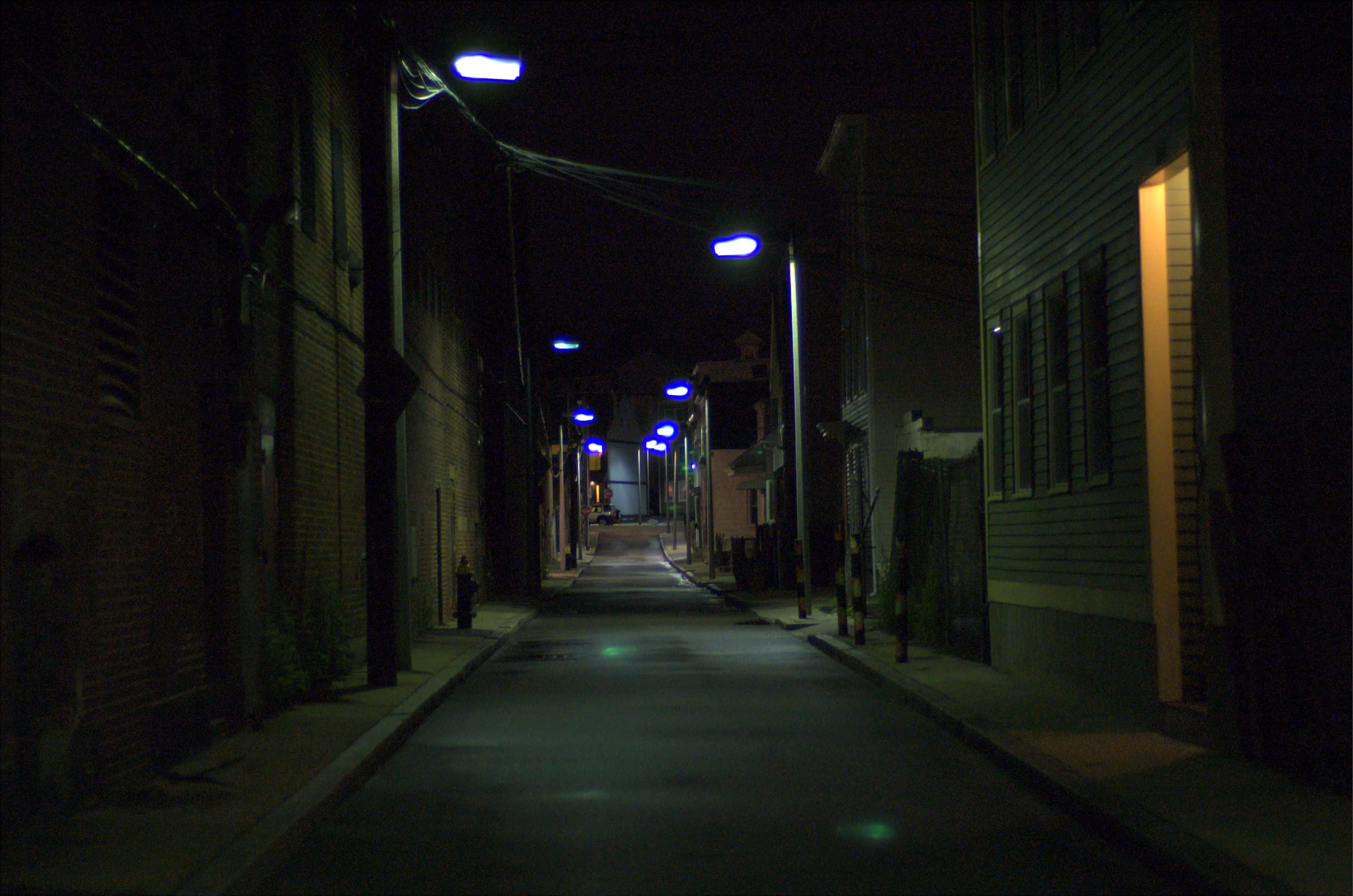 Alleyway 桌布 壁紙 — 夜晚一條狹窄的城市小巷，被藍色街燈照亮，在磚木建築之間投下長長的影子。