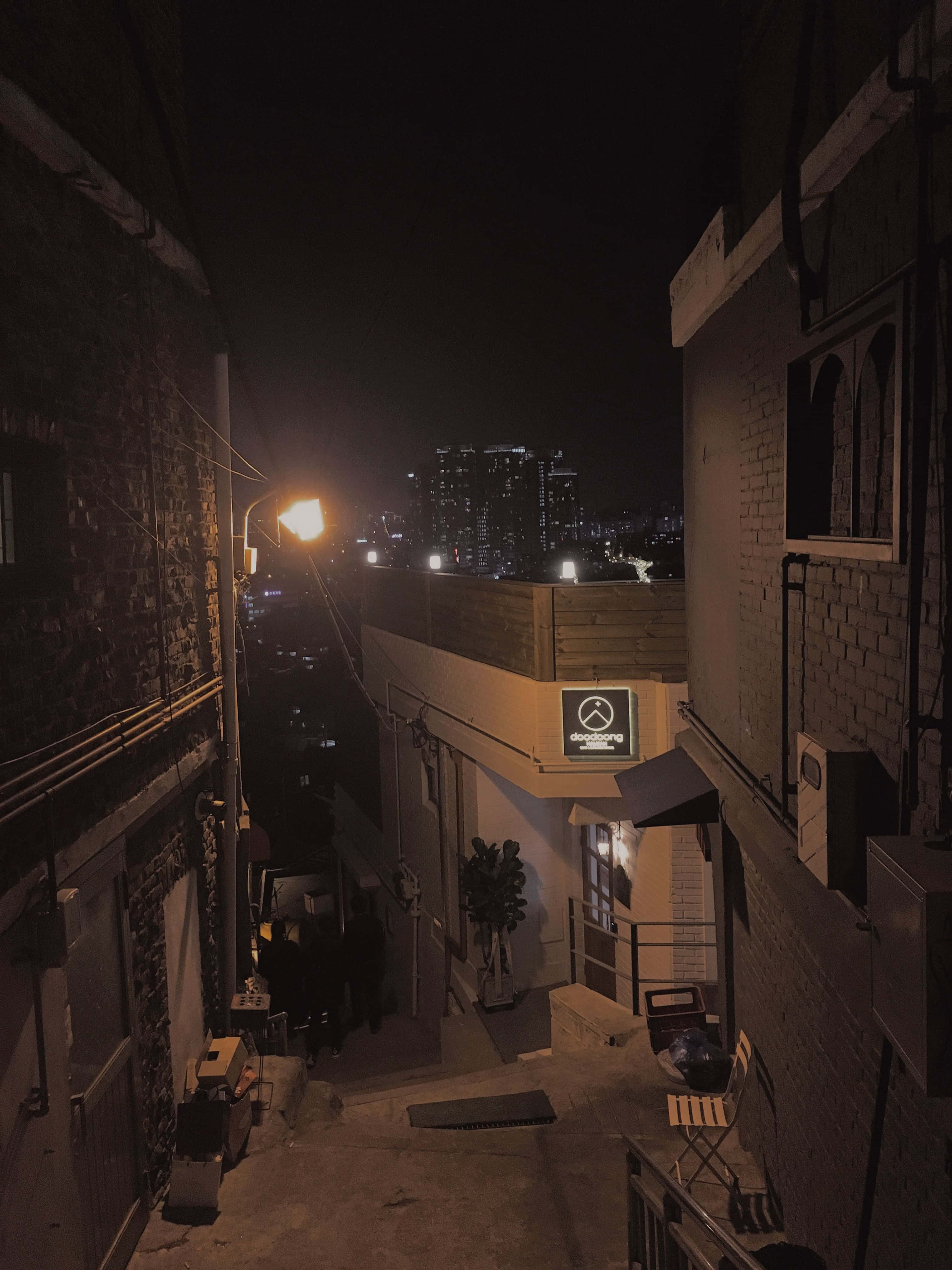 暗黑風格 桌布 壁紙 — 夜晚一條狹窄的磚砌小巷，由單一街燈照亮，遠處的城市天際線在背後閃耀。