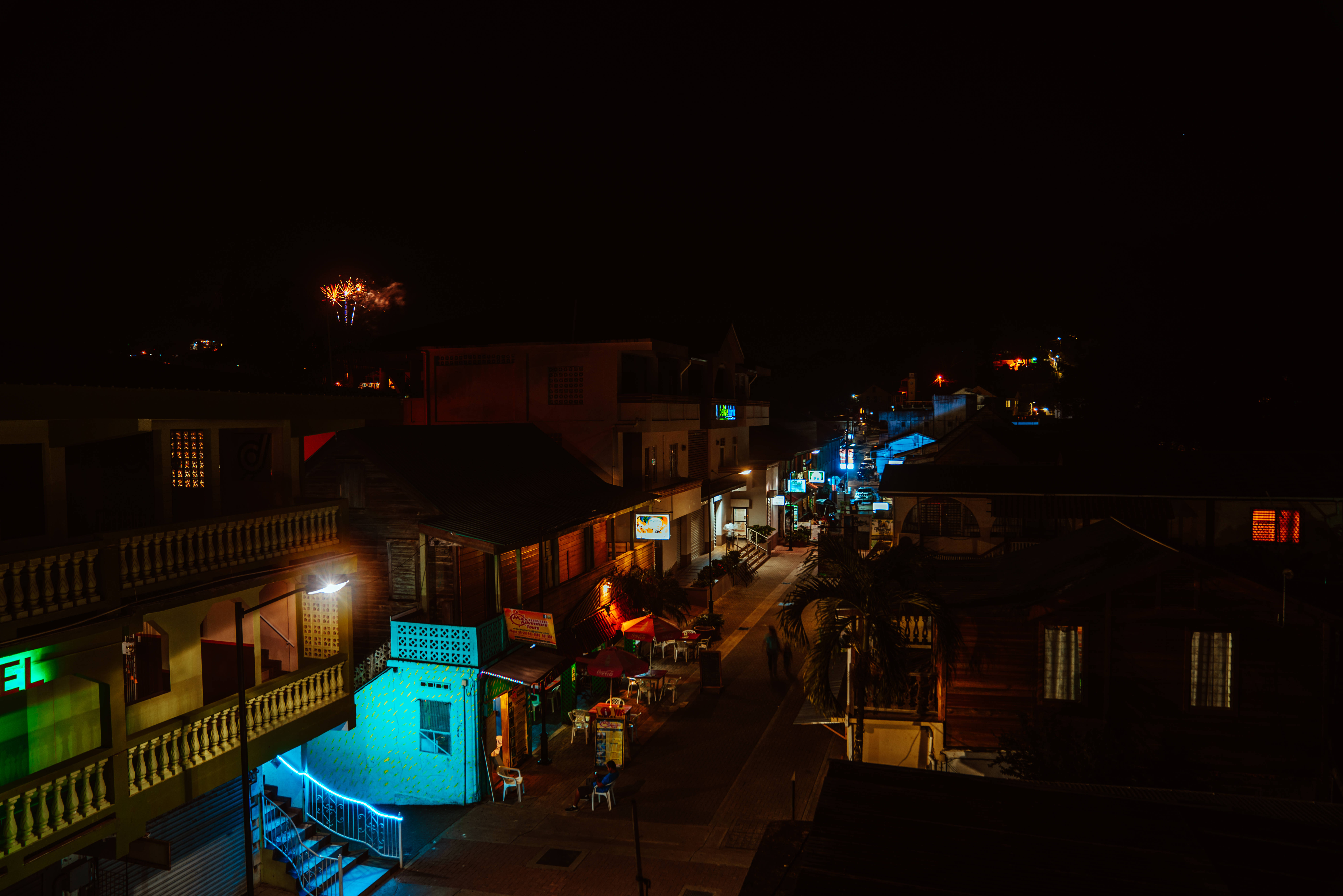 Belize 桌布 壁紙 — 夜晚一條狹窄的街道，被色彩繽紛的霓虹燈招牌和溫暖的路燈照亮，在建築物和棕櫚樹上投下長長的影子。