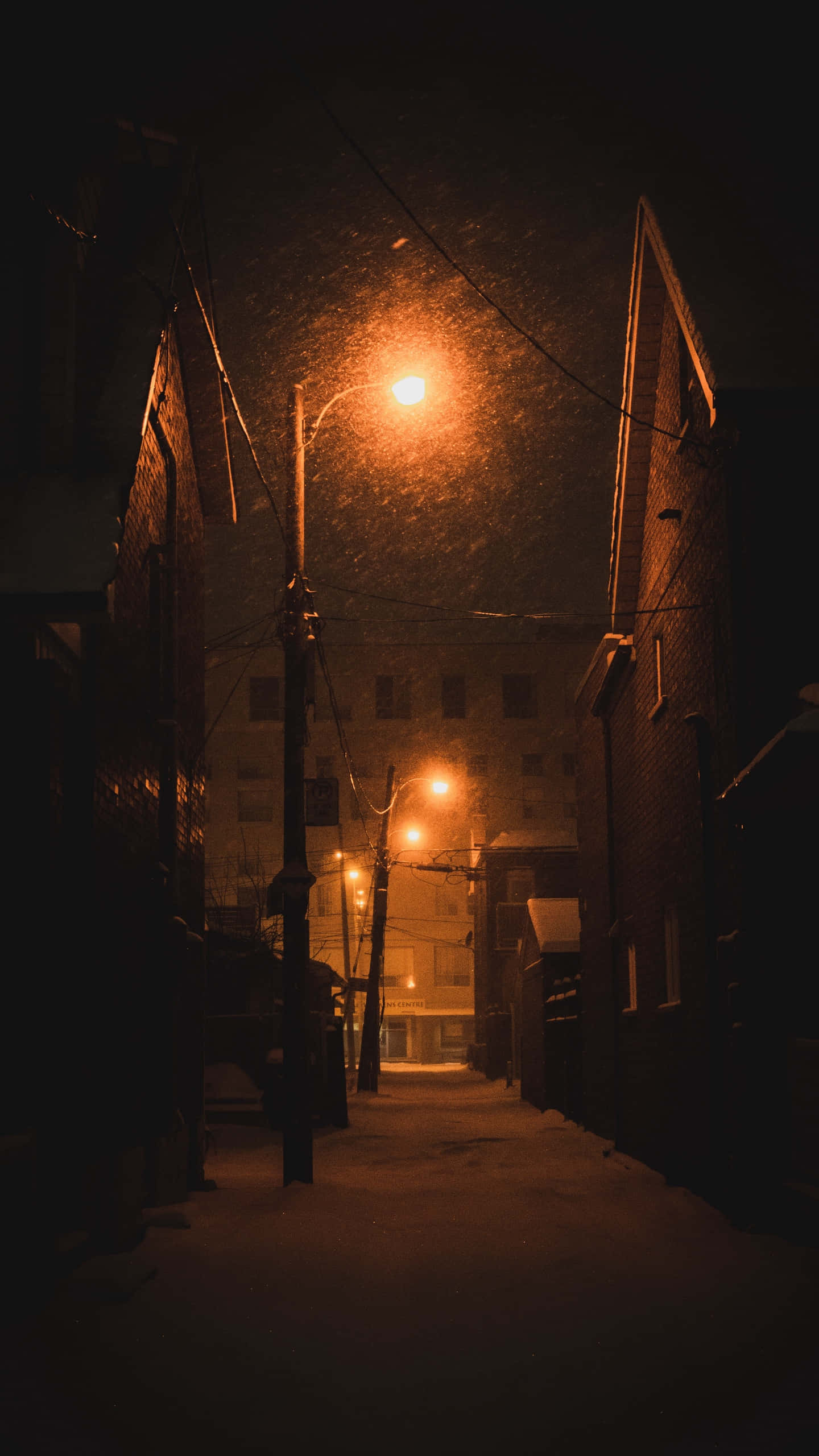 Street Light 桌布 壁紙 — 夜晚一條狹窄的雪巷，被溫暖的路燈照亮，在磚建築上投下長長的影子。