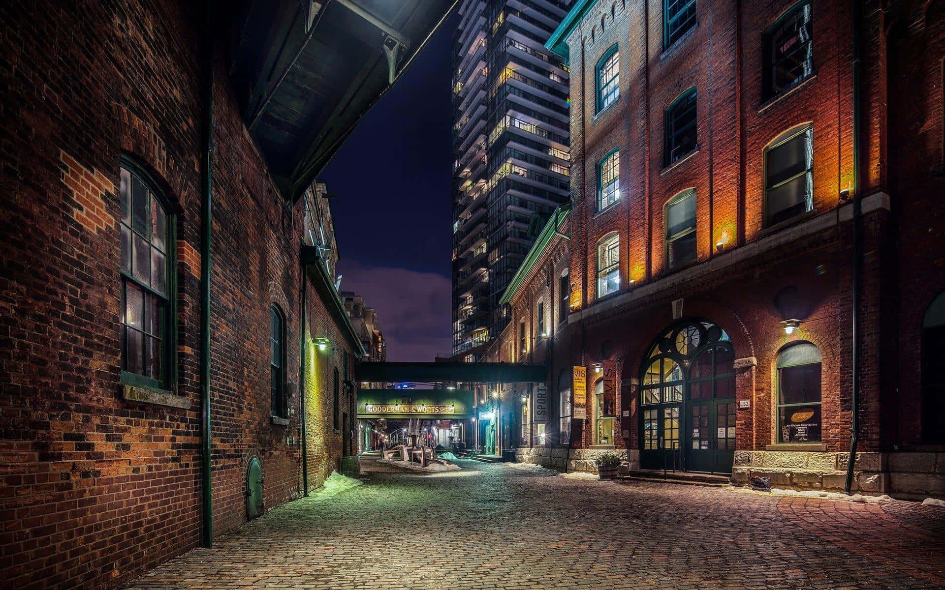 Alleyway 桌布 壁紙 — 夜晚一條狹窄的鵝卵石小巷，兩旁是歷史悠久的磚砌建築，溫暖的燈光與背景中高聳的現代摩天大樓形成對比。
