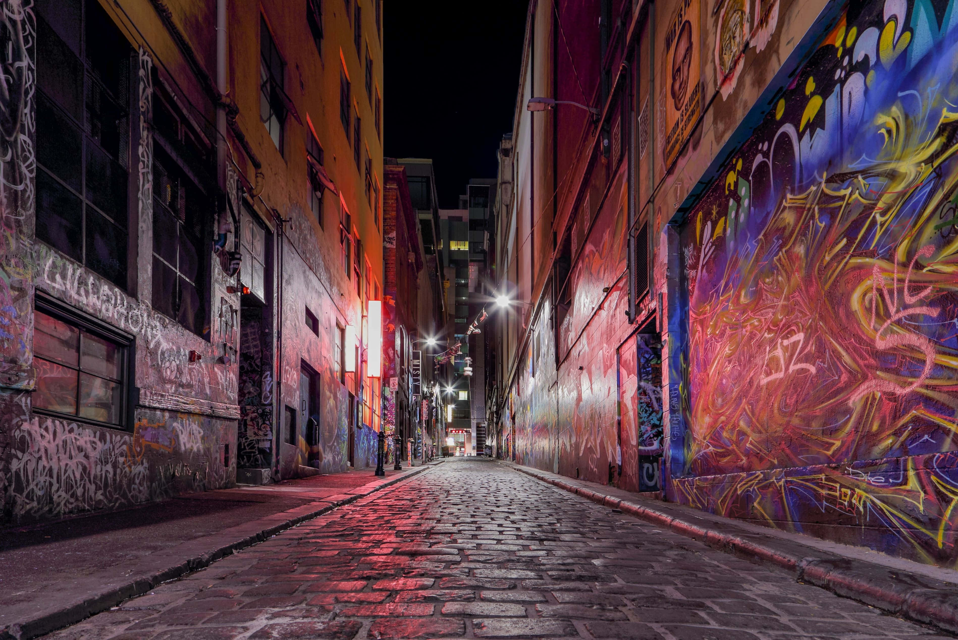Alleyway 桌布 壁紙 — 夜晚一條狹窄的鵝卵石小巷，被充滿活力的塗鴉壁畫和街燈照亮，在風化的牆壁上投射出紅色和藍色的色調。