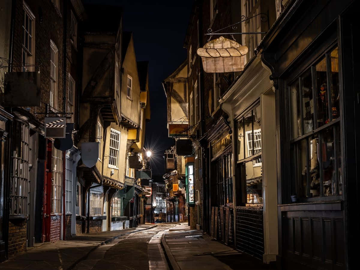York 桌布 壁紙 — 夜晚一條狹窄的鵝卵石小巷，被歷史悠久的歐洲建築中溫暖的燈籠和商店櫥窗照亮。