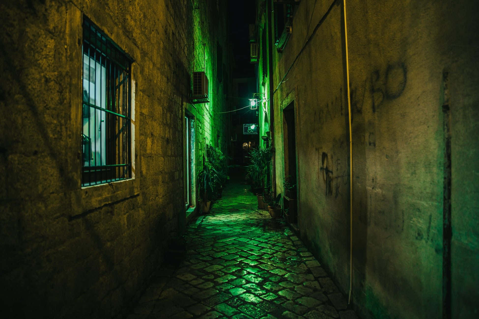 Alleyway 桌布 壁紙 — 夜晚一條狹窄的鵝卵石小巷，被遠處招牌散發的詭異綠色霓虹燈光籠罩，在風化的石牆上投下長長的陰影。