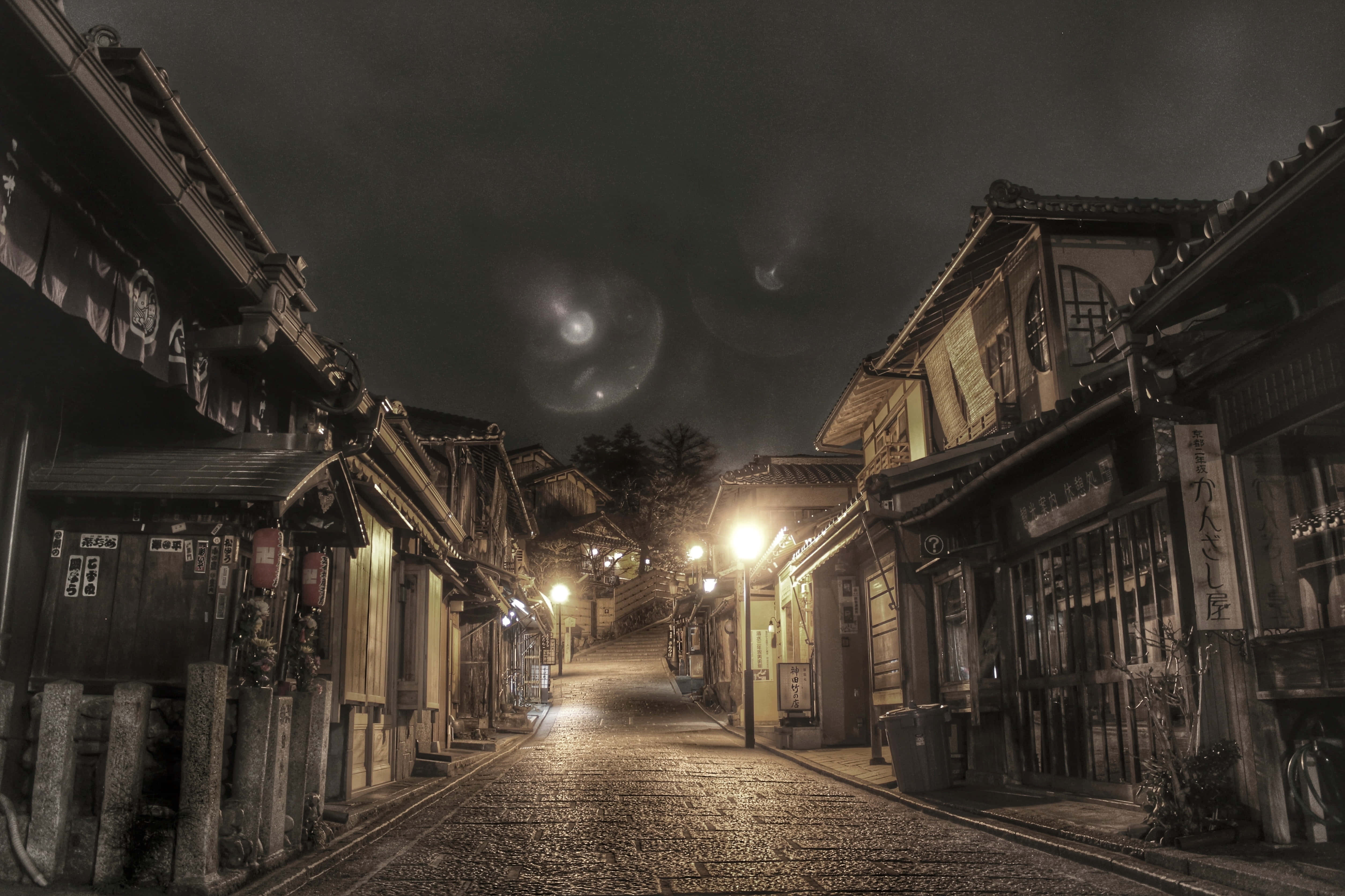 桌布 壁紙 — 夜晚一條狹窄的鵝卵石街道，兩旁是傳統木建築，在黑暗的天空下帶有鏡頭光暈。